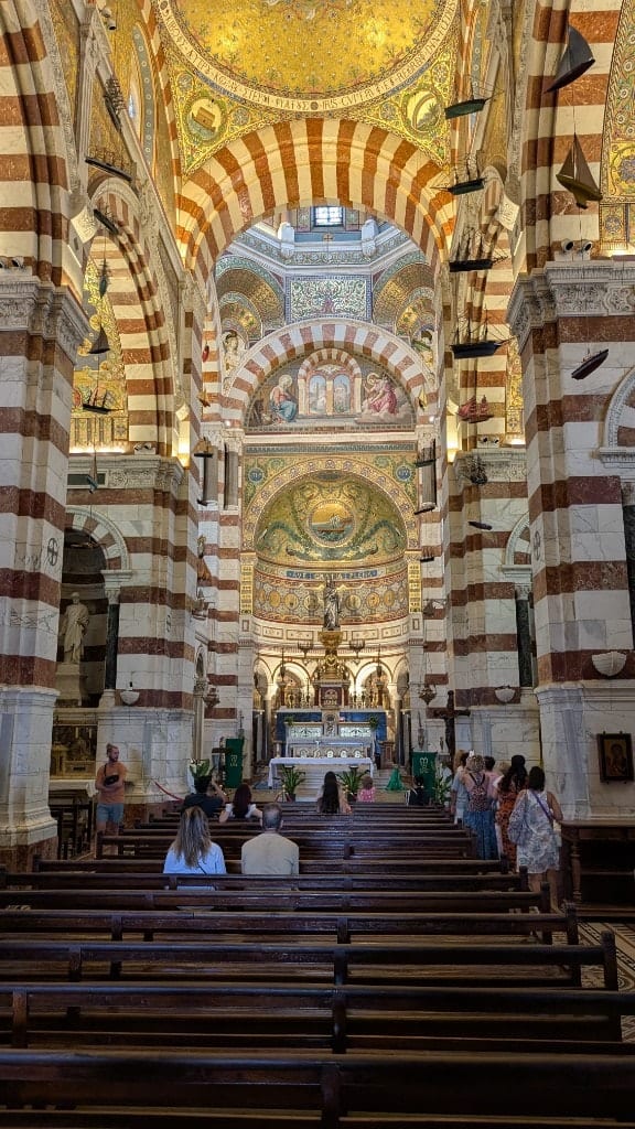 The interior of Notre-Dame de la Garde with red-and-white striped stone arches, a gold mosaic ceiling, model ships hanging overhead, and visitors in the pews