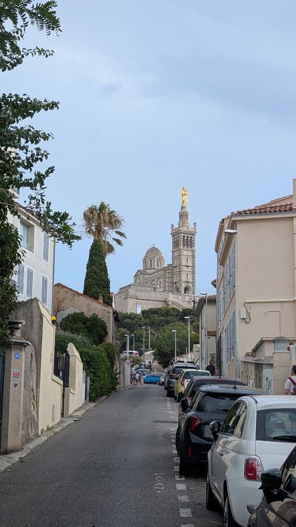 A quiet residential street climbing toward the basilica of Notre-Dame de la Garde, its golden Madonna catching the light above parked cars, a palm and a cypress
