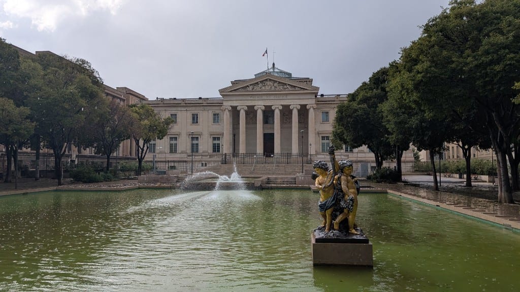 The neoclassical Palais de Justice courthouse in Marseille behind a green reflecting pool with a fountain and a gilded cherub statue, framed by plane trees