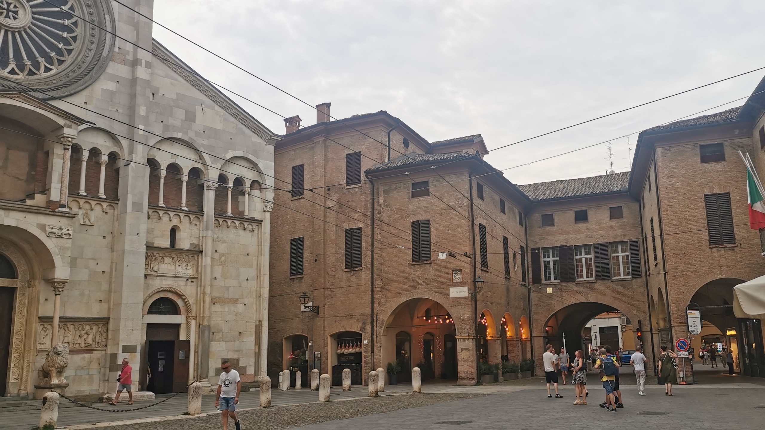 The carved stone front of Modena Cathedral with its rose window and a stone lion by the door, beside brick arcades on Piazza Duomo with hams hanging in a shop window