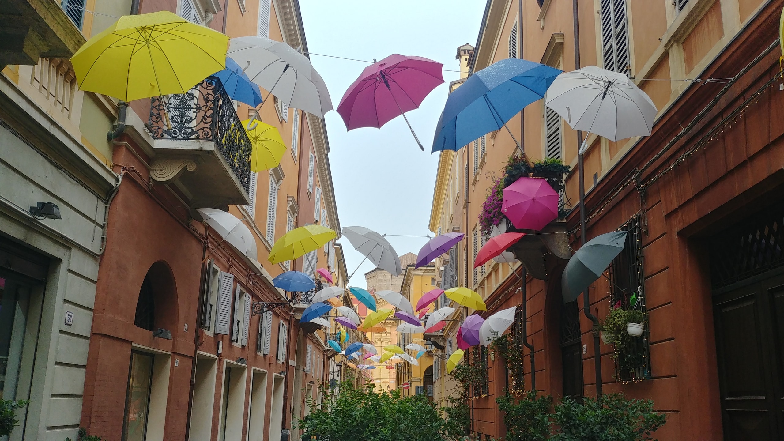 Dozens of open umbrellas in yellow, pink, blue, and white hanging on wires above a narrow street of orange and red houses in Modena