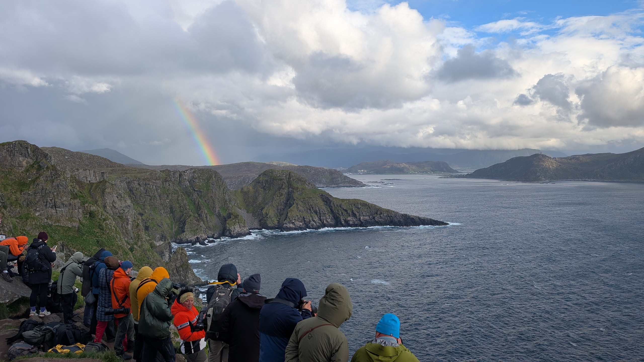 People in colorful outdoor jackets lined along a rocky coastal cliff with a rainbow over deep blue water