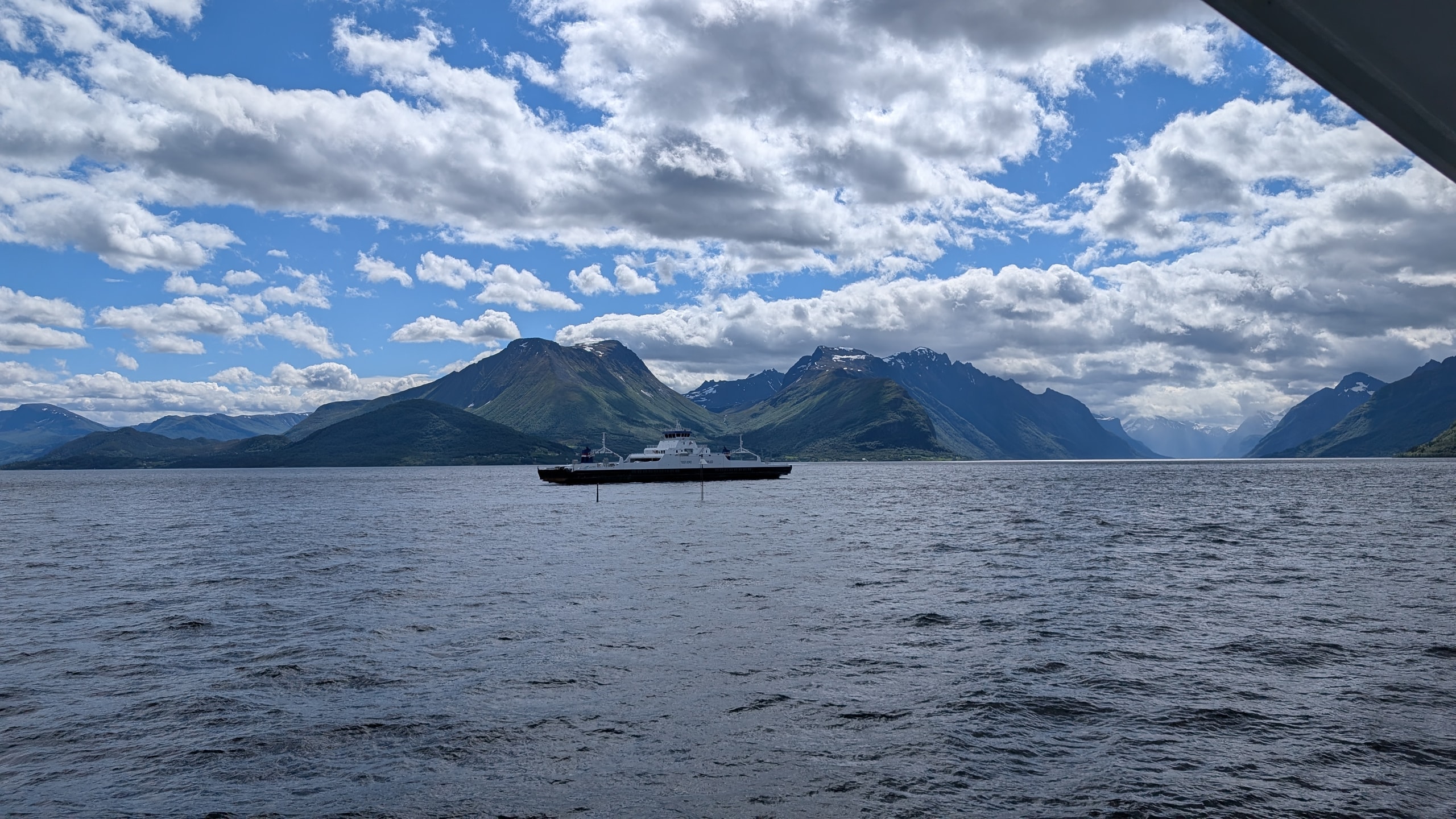 A white car ferry crossing a blue fjord with steep green mountains and lingering snow on the ridges