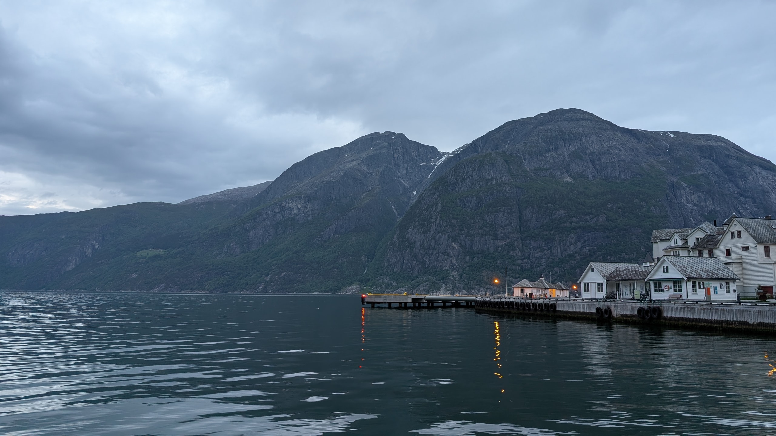 White wooden buildings on a fjord pier with warm window light reflecting in calm water under steep dark mountains