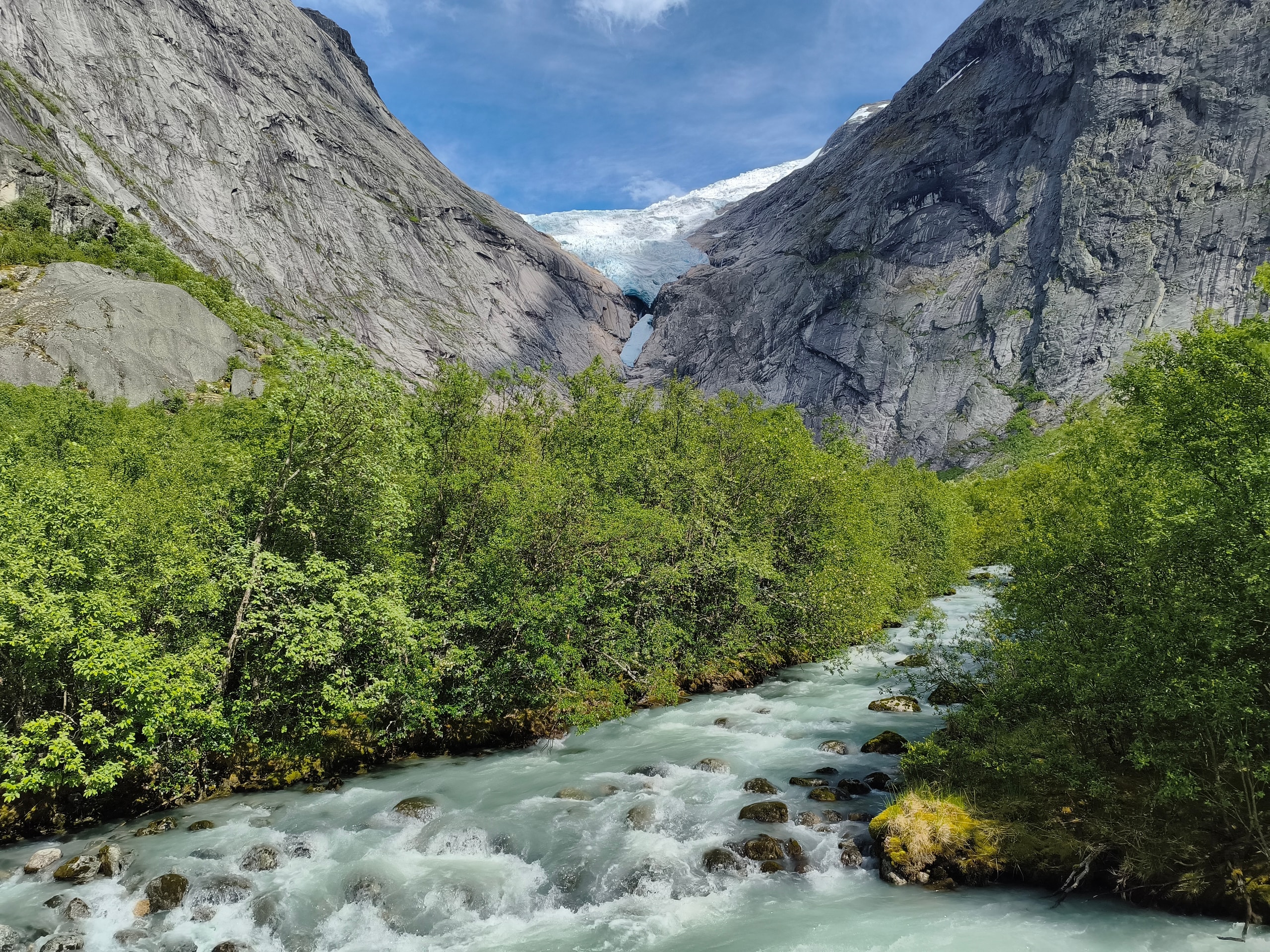 A milky glacial river rushing toward the camera with a white glacier between steep grey mountain walls