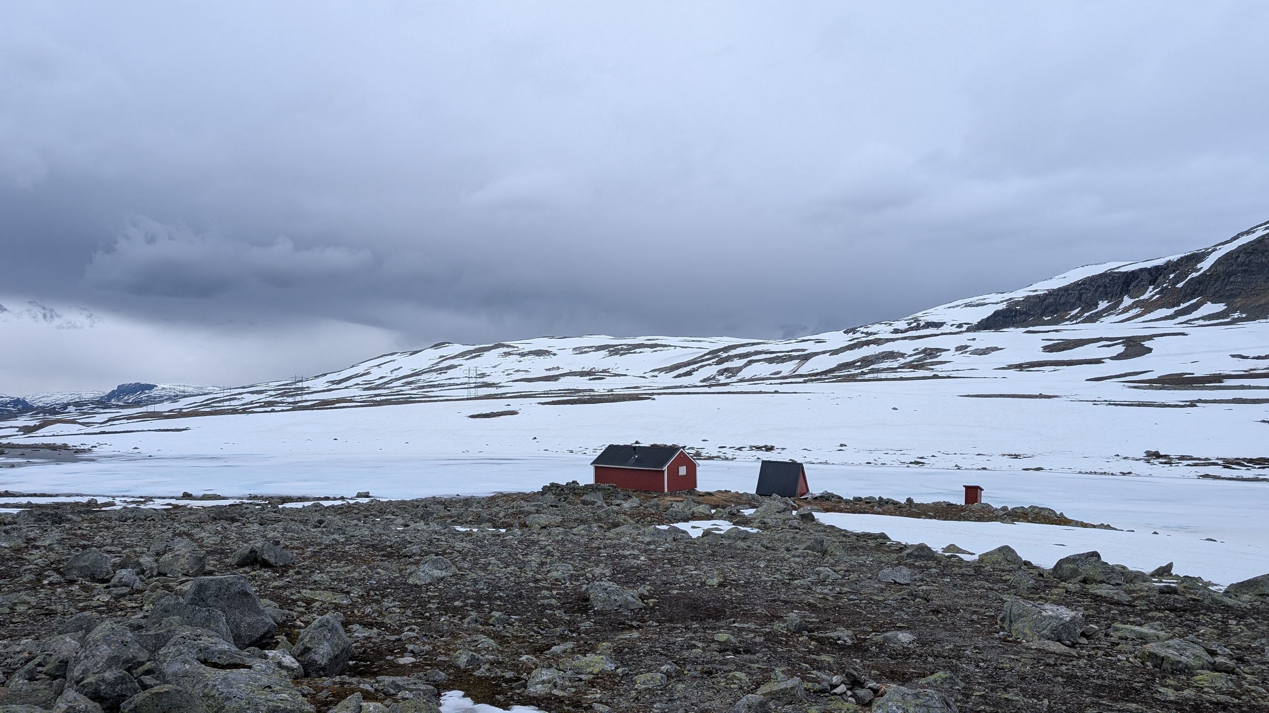 A small red wooden mountain cabin on rocky snowy ground with snow-covered hills under a grey sky