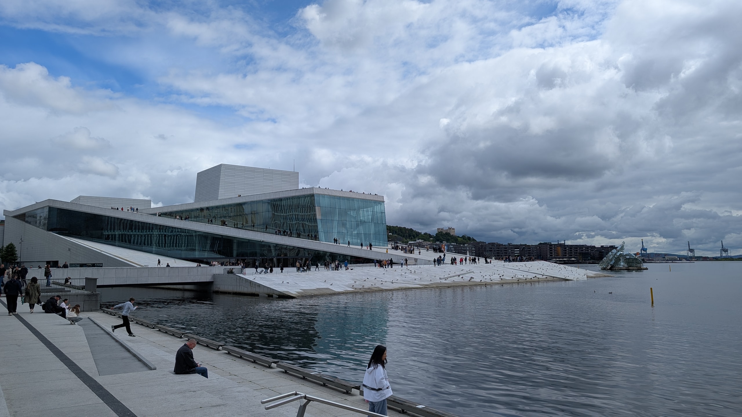 Oslo Opera House rising from the harbour with people walking the white sloping roof under mixed cloud