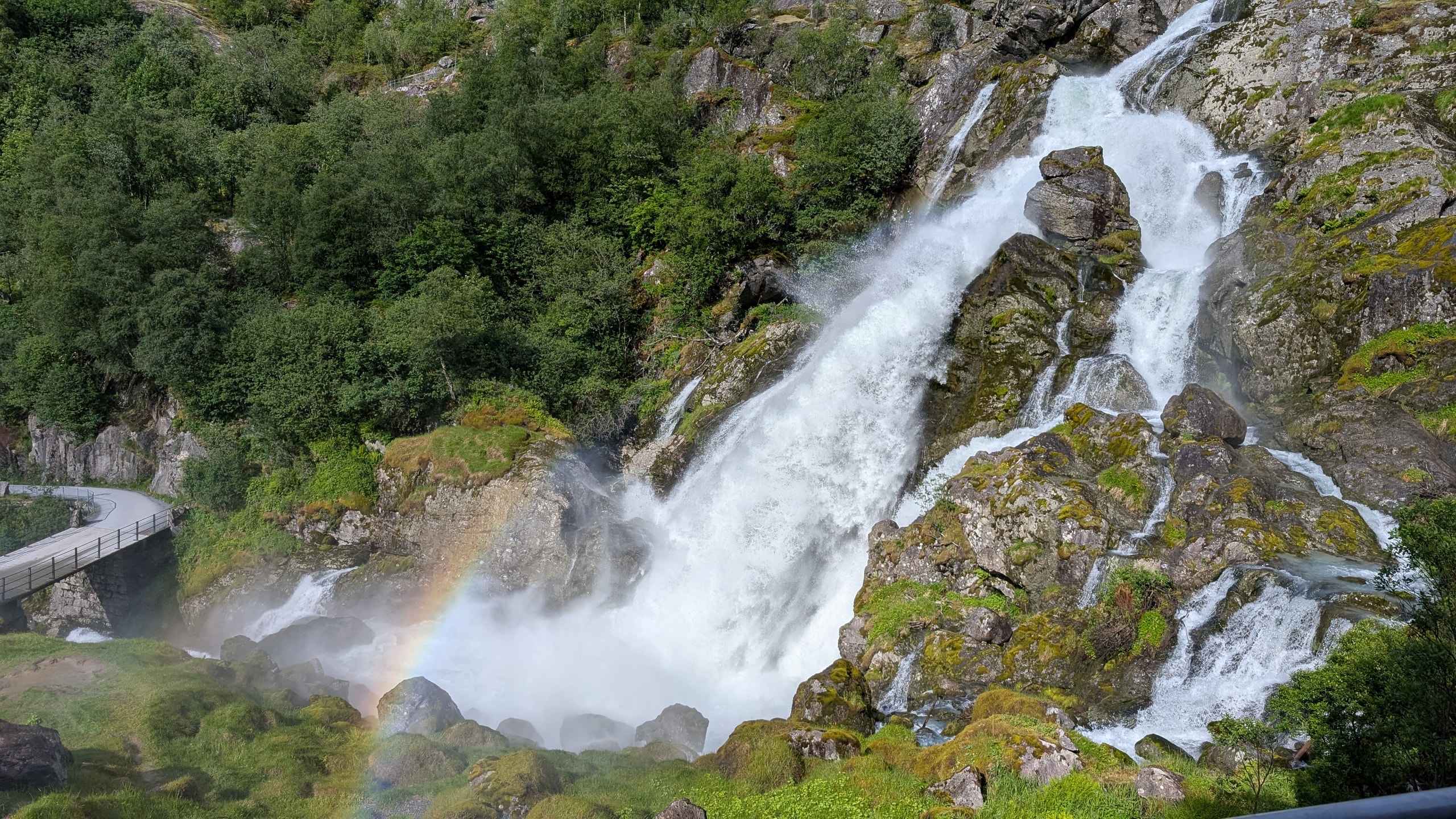 A multi-tiered waterfall crashing down mossy rock with a bright rainbow in the spray beside a road railing
