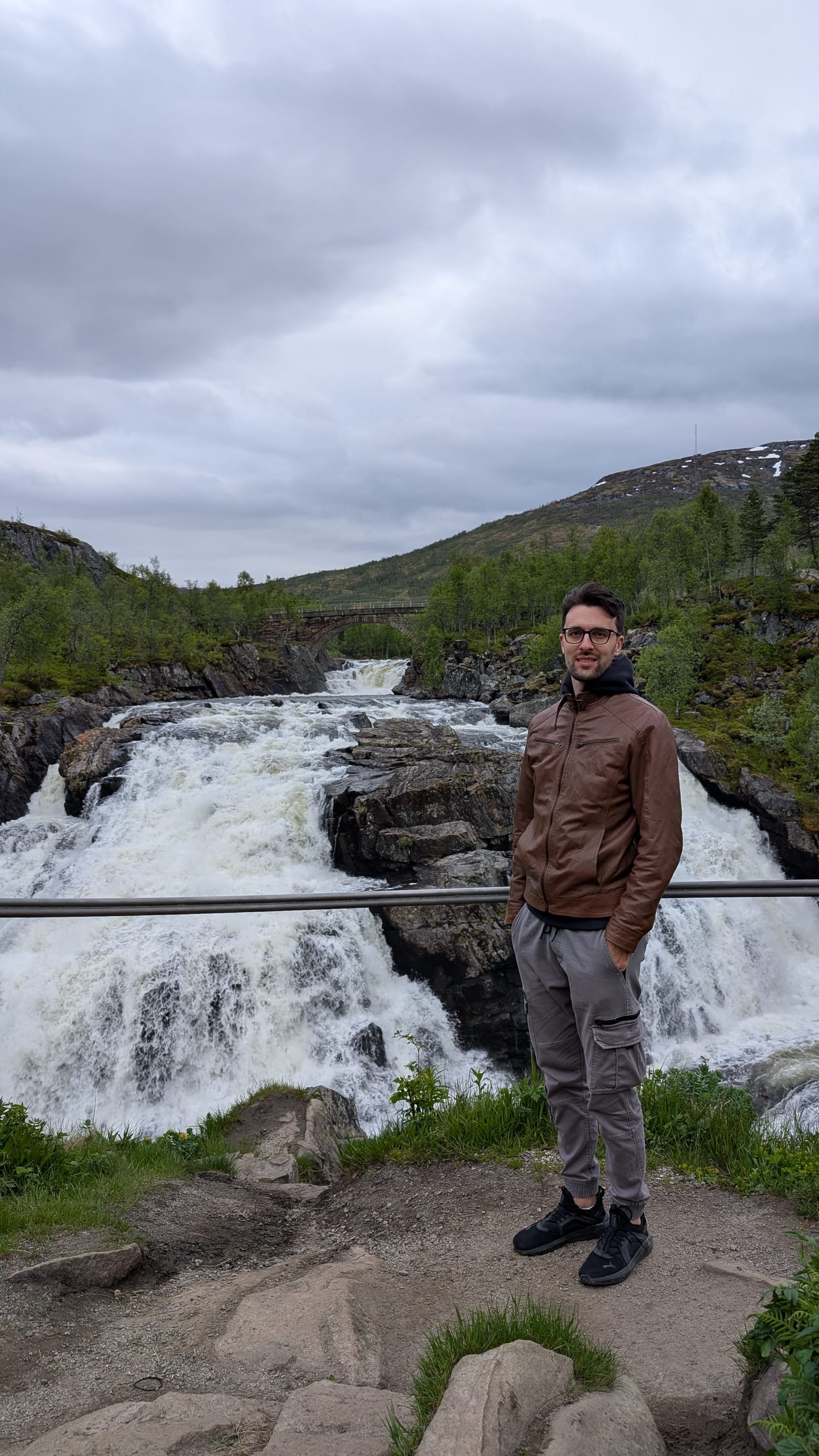 Erik wearing a brown leather jacket beside a wide waterfall and stone bridge in western Norway
