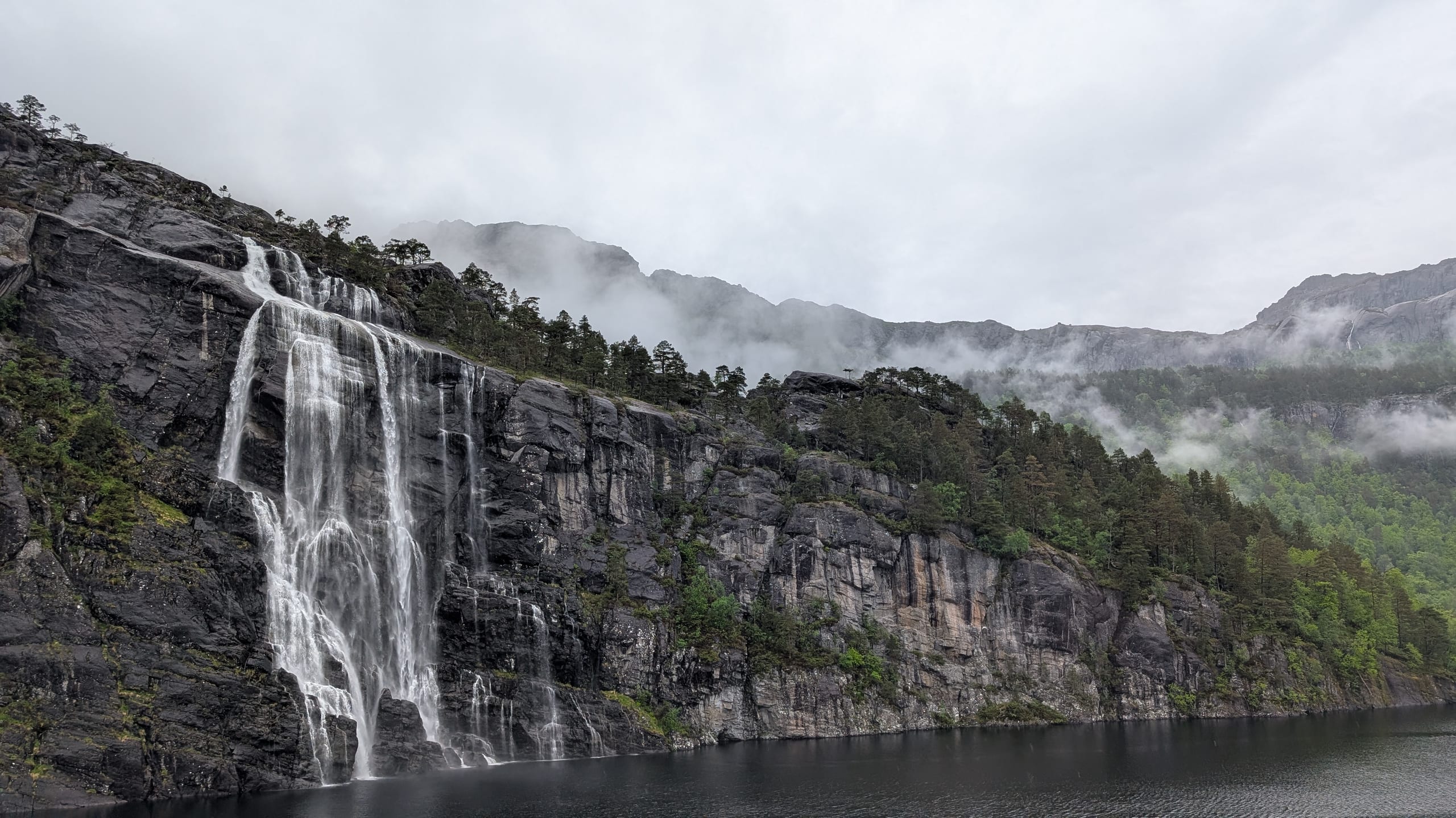 A tall waterfall cascading down a rock face beside a Norwegian road with green slopes and mist
