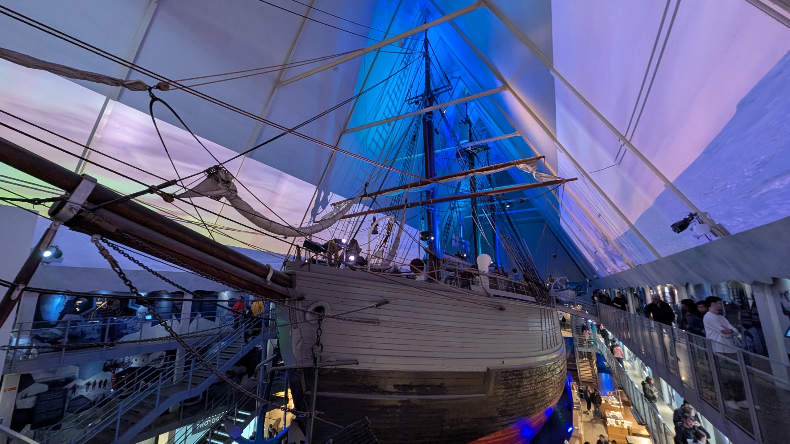 The wooden polar ship Fram inside its A-frame museum hall in Oslo, lit in blue with visitors on surrounding walkways
