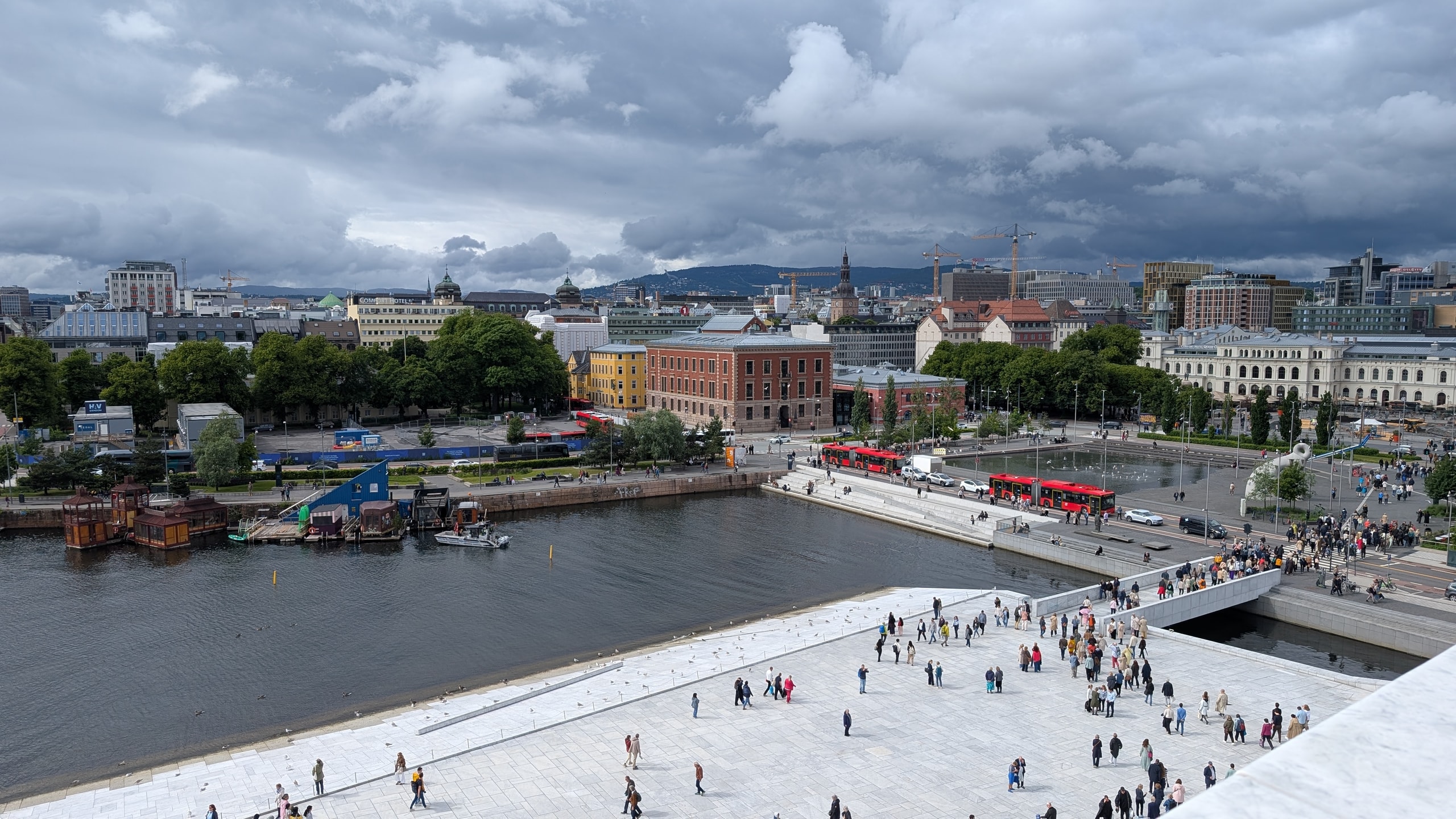 View across the white marble Opera House roof toward Oslo’s harbour, city buses, and forested hills