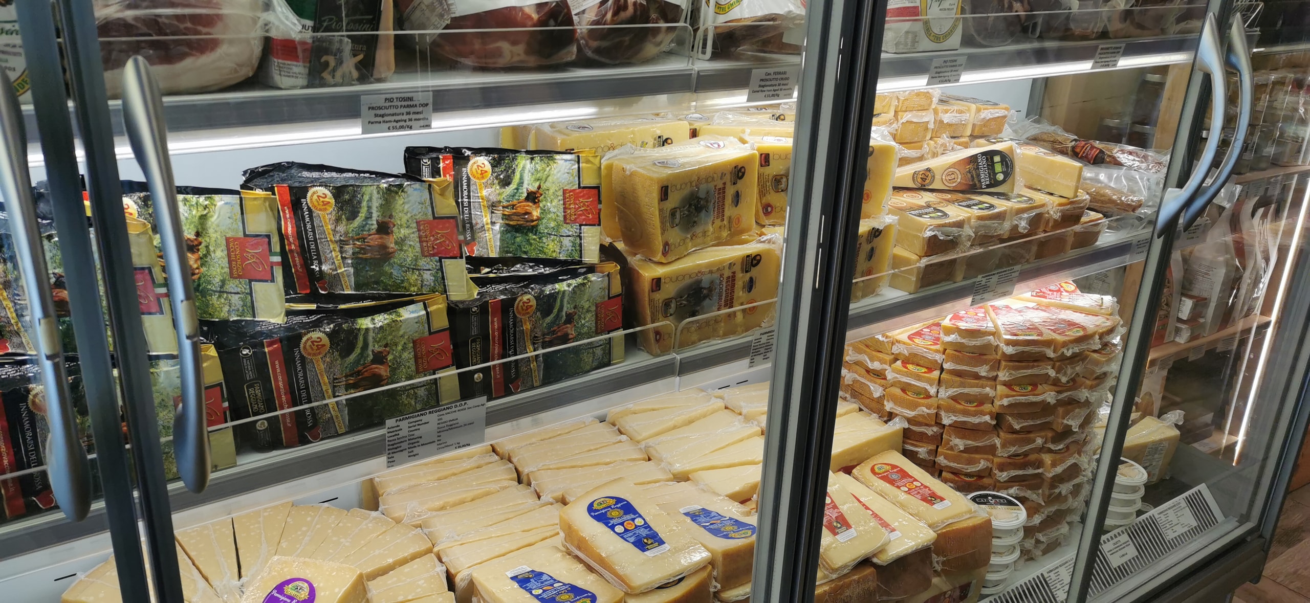 A glass-fronted deli fridge in Parma stacked with vacuum-packed Parmigiano Reggiano wedges and prosciutto, with printed price labels on the shelves