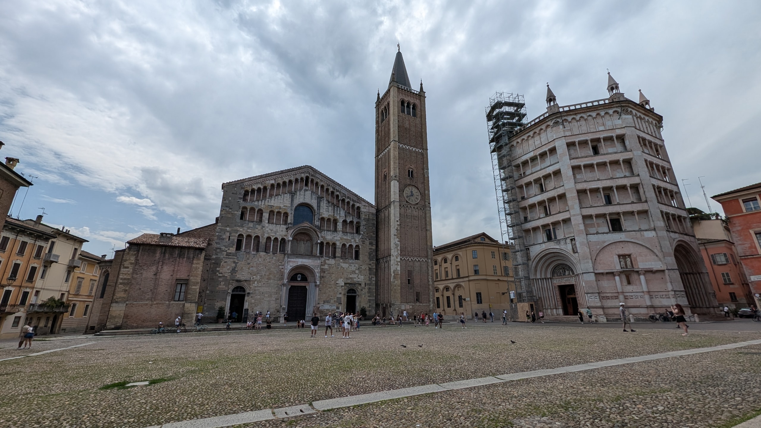 Parma's Romanesque cathedral, its brick bell tower, and the pink marble octagonal baptistery partly under scaffolding on a wide cobbled square under grey clouds