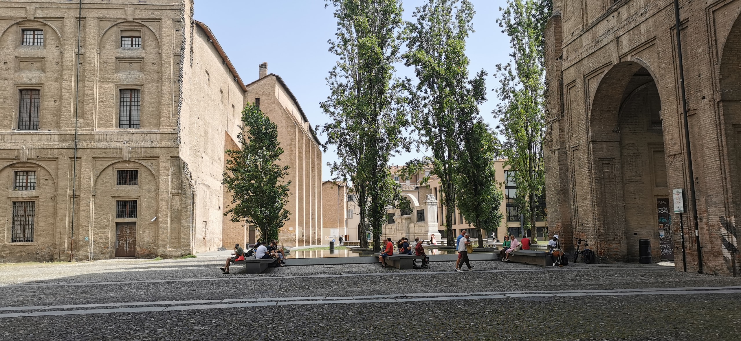 The bare brick walls and tall open arches of Parma's Palazzo della Pilotta beside Piazzale della Pace, with poplar trees, a shallow pool, and people sitting on stone benches