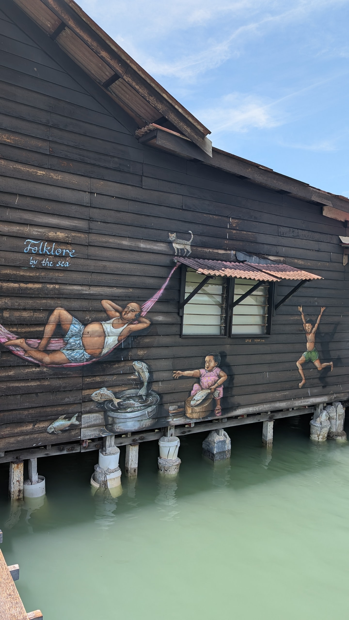 Mural of daily jetty life painted on dark wooden stilthouse siding above green water at a George Town clan jetty