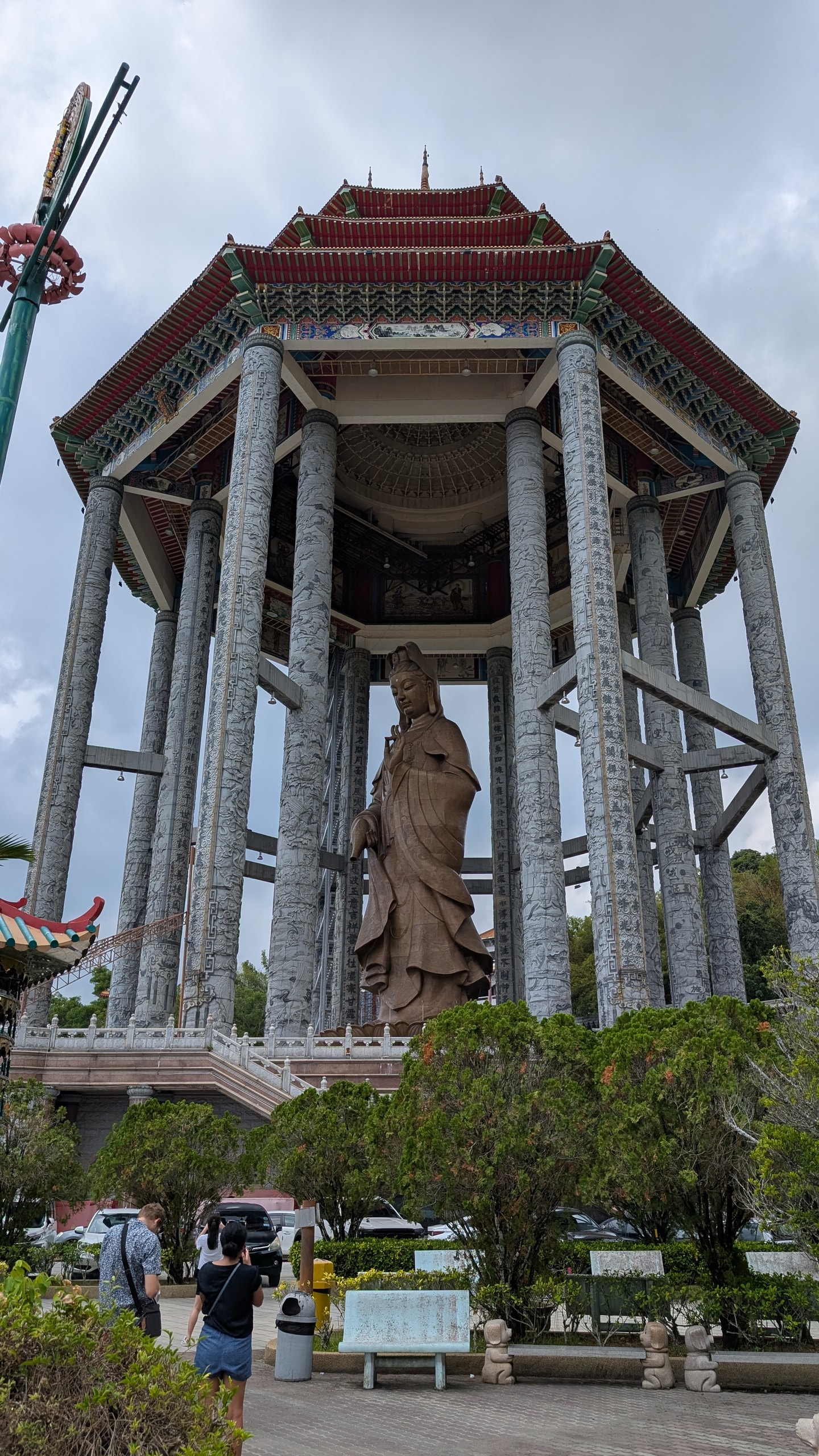 Massive bronze Guan Yin statue under an octagonal pavilion with carved stone columns at Kek Lok Si, with visitors on the courtyard below
