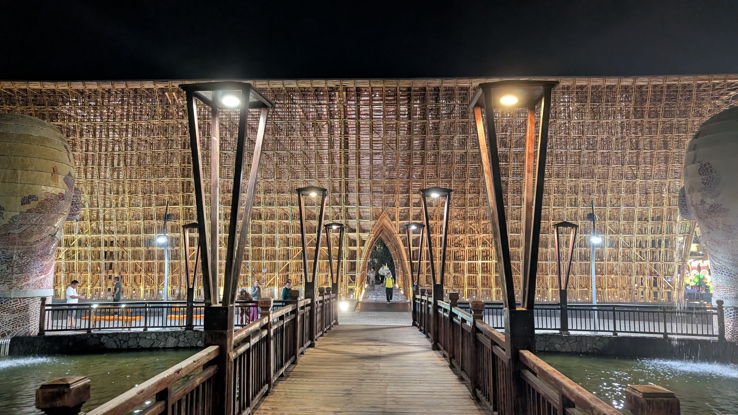 A large illuminated bamboo hall at night with a pointed arch entrance, seen from a wooden footbridge over dark water