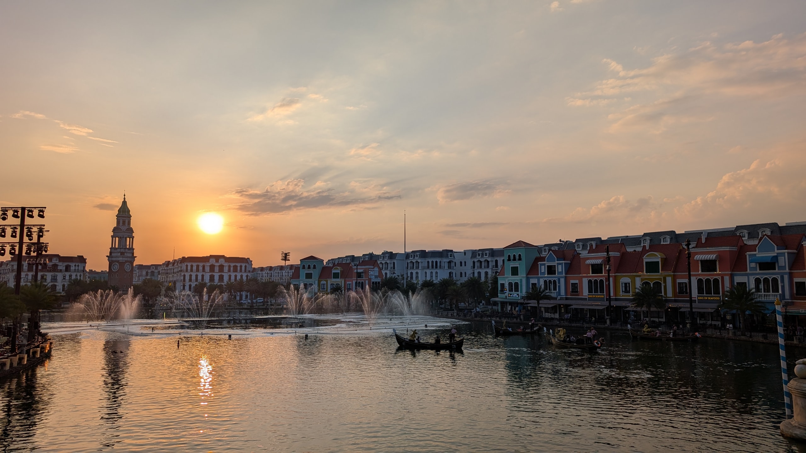 Fountains spraying across a lake at sunset with a clock tower on the left and rows of colourful buildings on the right