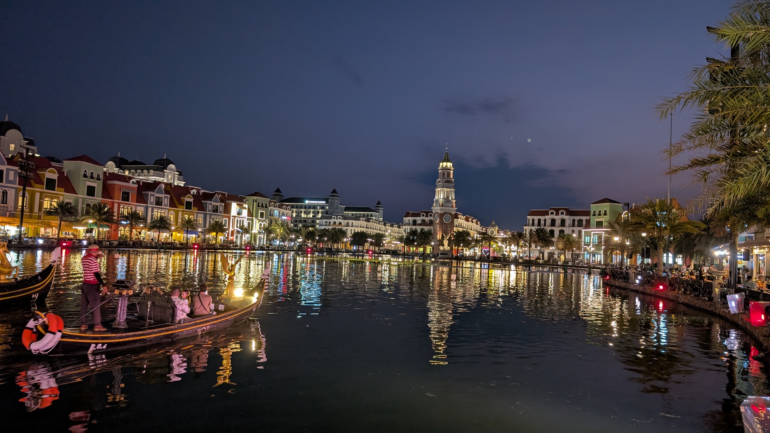 A gondolier poling a boat with passengers across a lit lake at night, with illuminated buildings and a clock tower reflected in the water