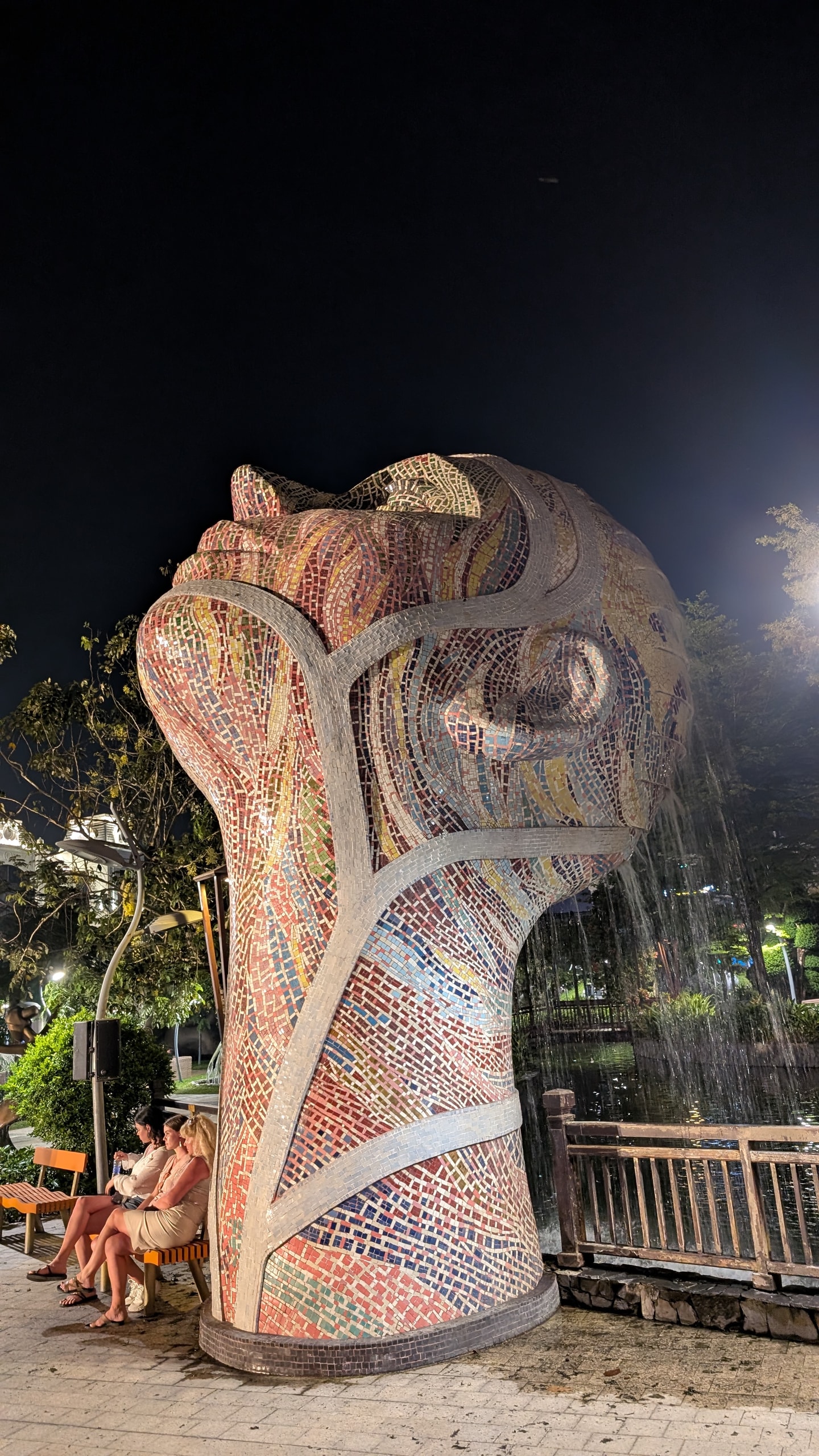 A tall mosaic-tiled sculpture of a human head lit at night, with two people sitting on a bench at its base for scale