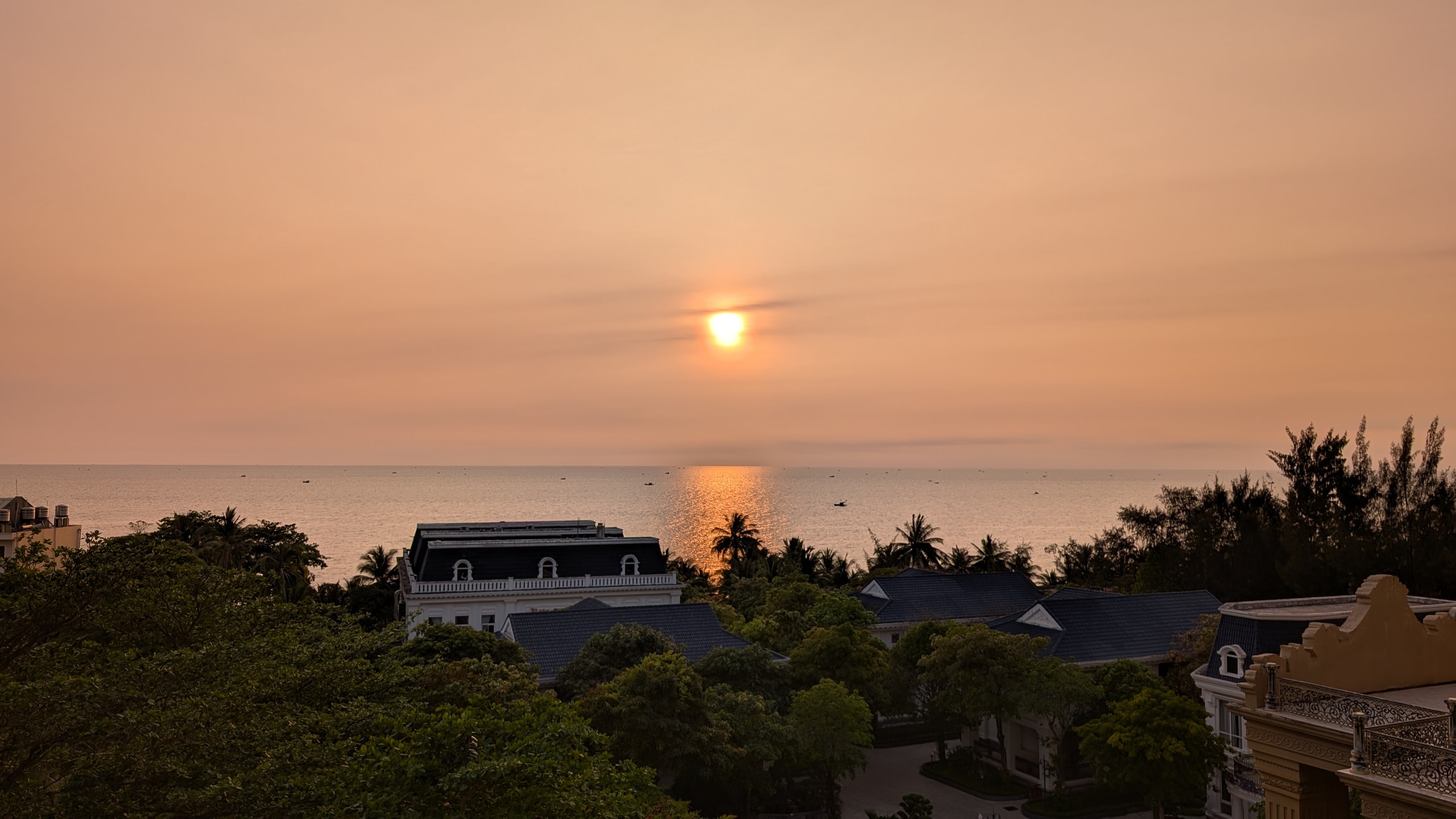 A hazy orange sunset over the sea with a gold path of light on the water, palm trees and resort roofs below