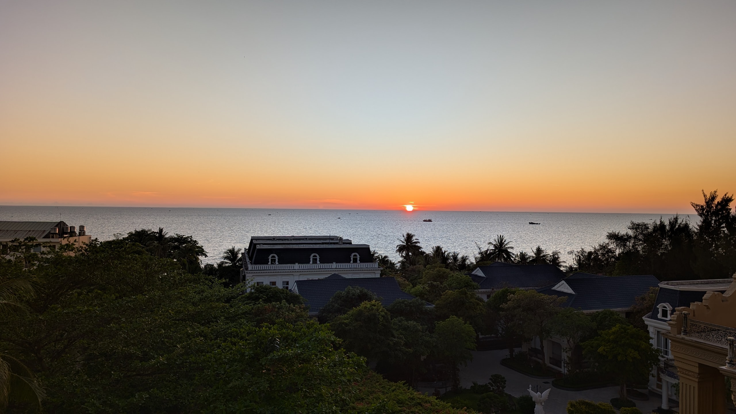 The sun sitting just above the horizon over the Gulf of Thailand, with resort roofs and palms dark in the foreground