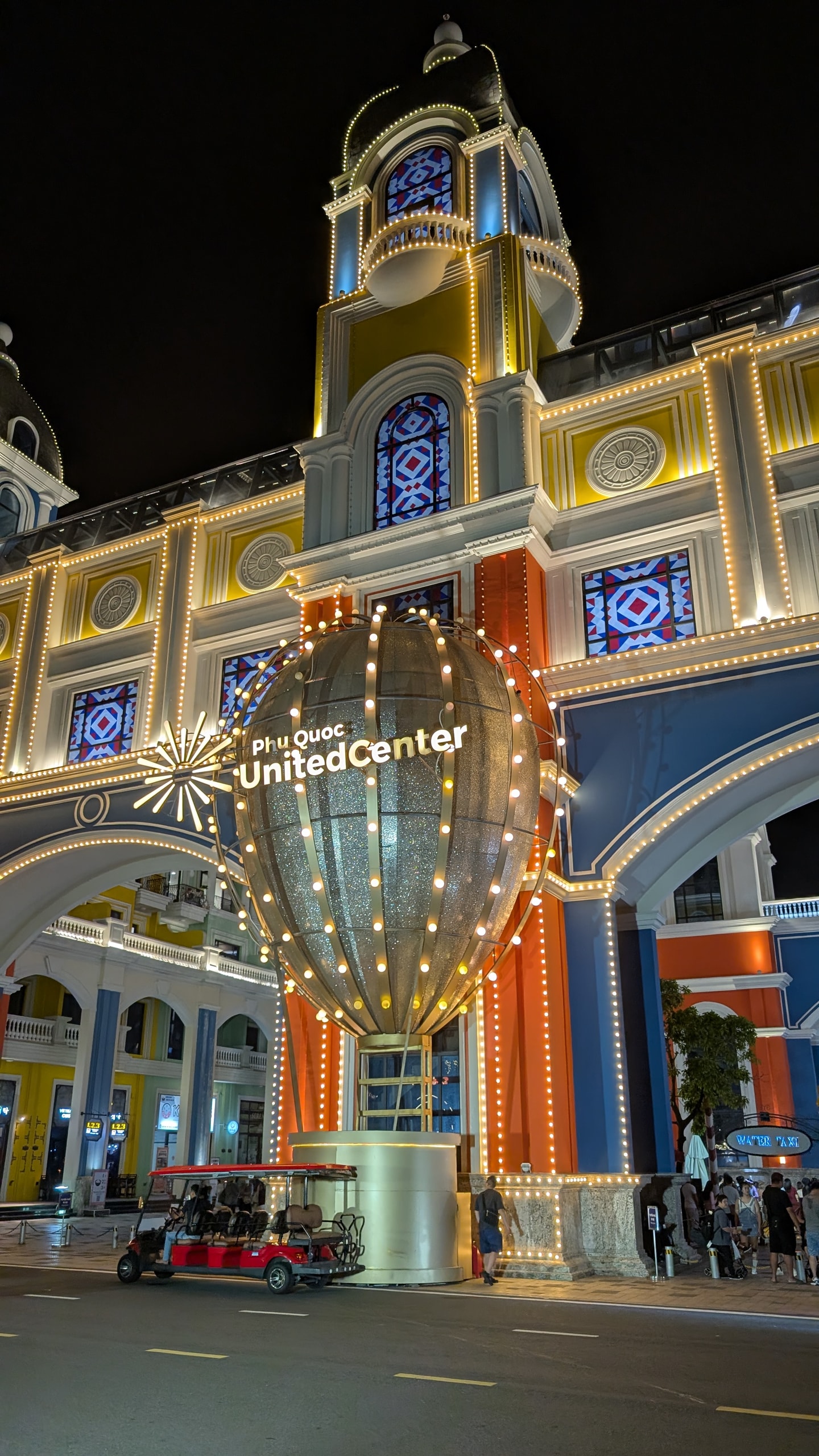 The illuminated entrance arch of Phu Quoc United Center at night with a lit balloon sculpture and an electric shuttle cart parked in front