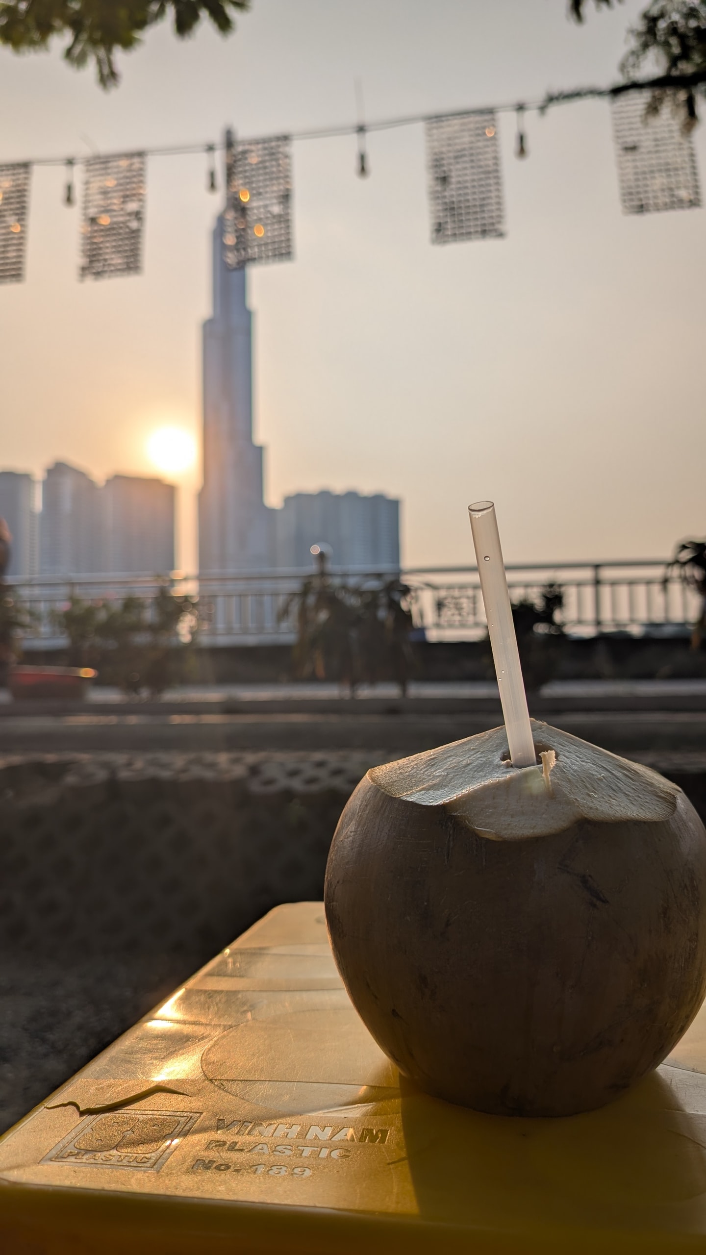 A fresh coconut with a straw on a yellow plastic table, Landmark 81 blurred in the sunset behind a metal railing