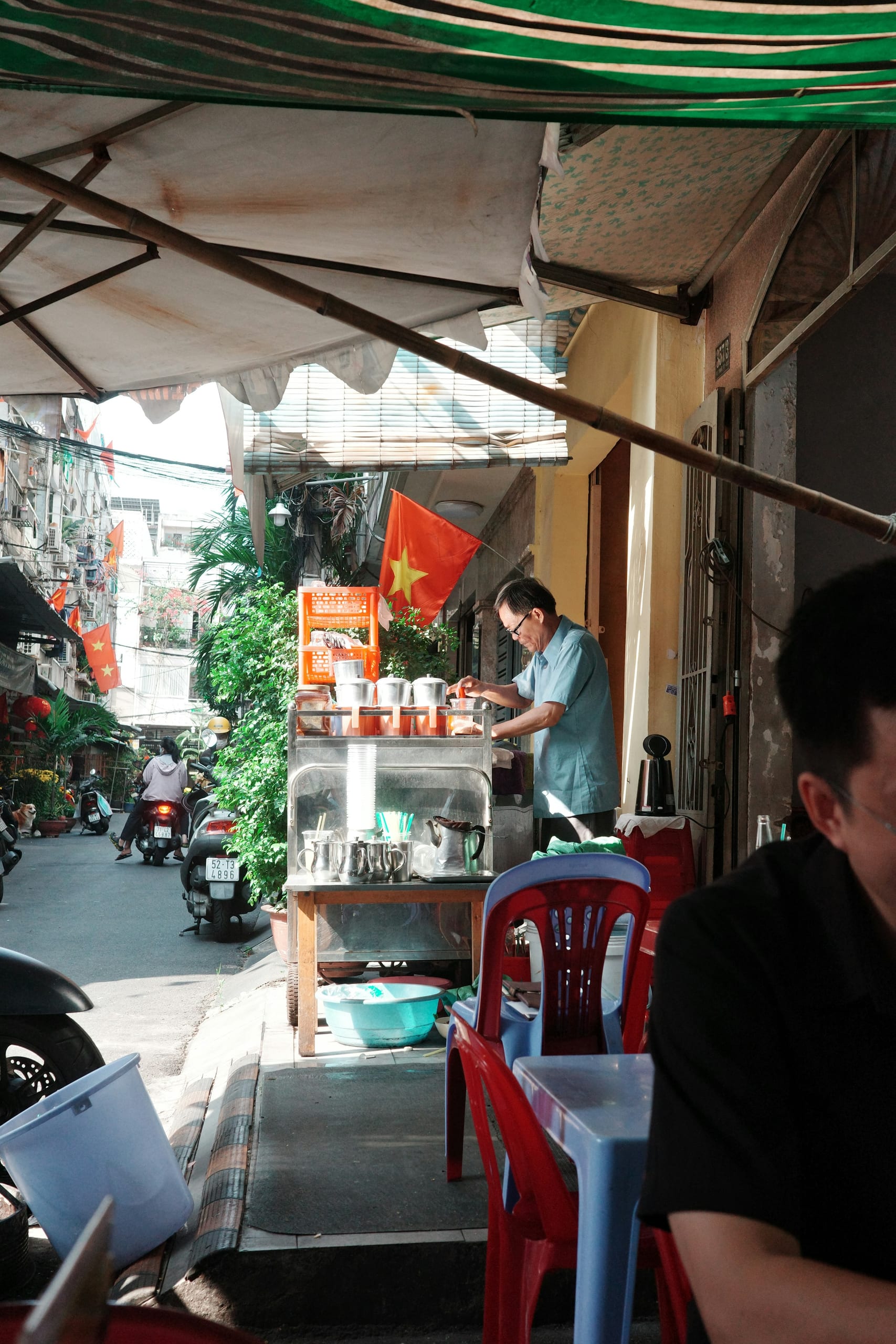 A street stall with plastic stools and a vendor preparing drinks under an awning in a Saigon alley