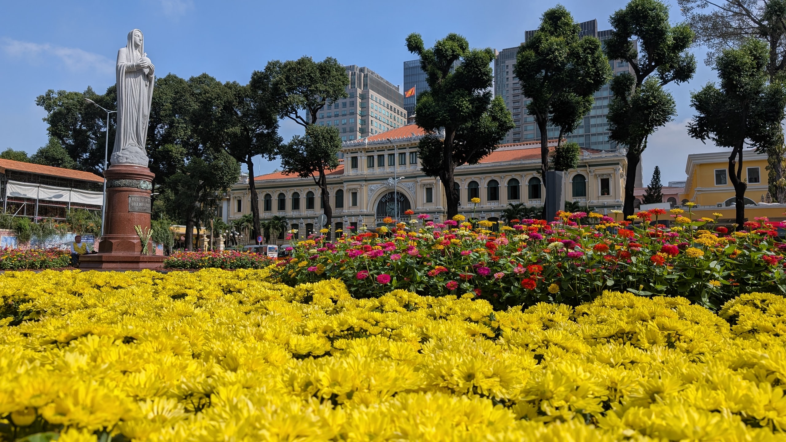Saigon Central Post Office behind yellow chrysanthemums and pink flower beds, with the Virgin Mary statue on the left