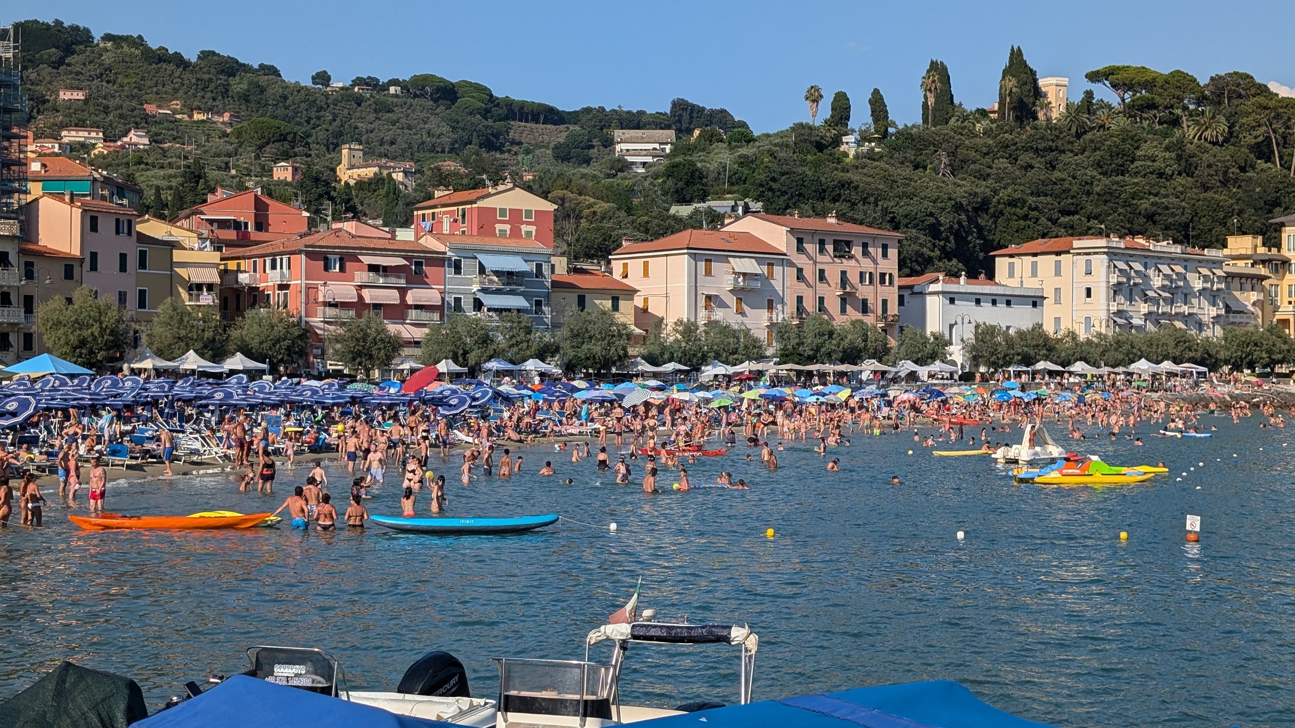 A crowded beach at San Terenzo packed with blue umbrellas, swimmers, and paddleboards in front of pastel seafront buildings and green hills