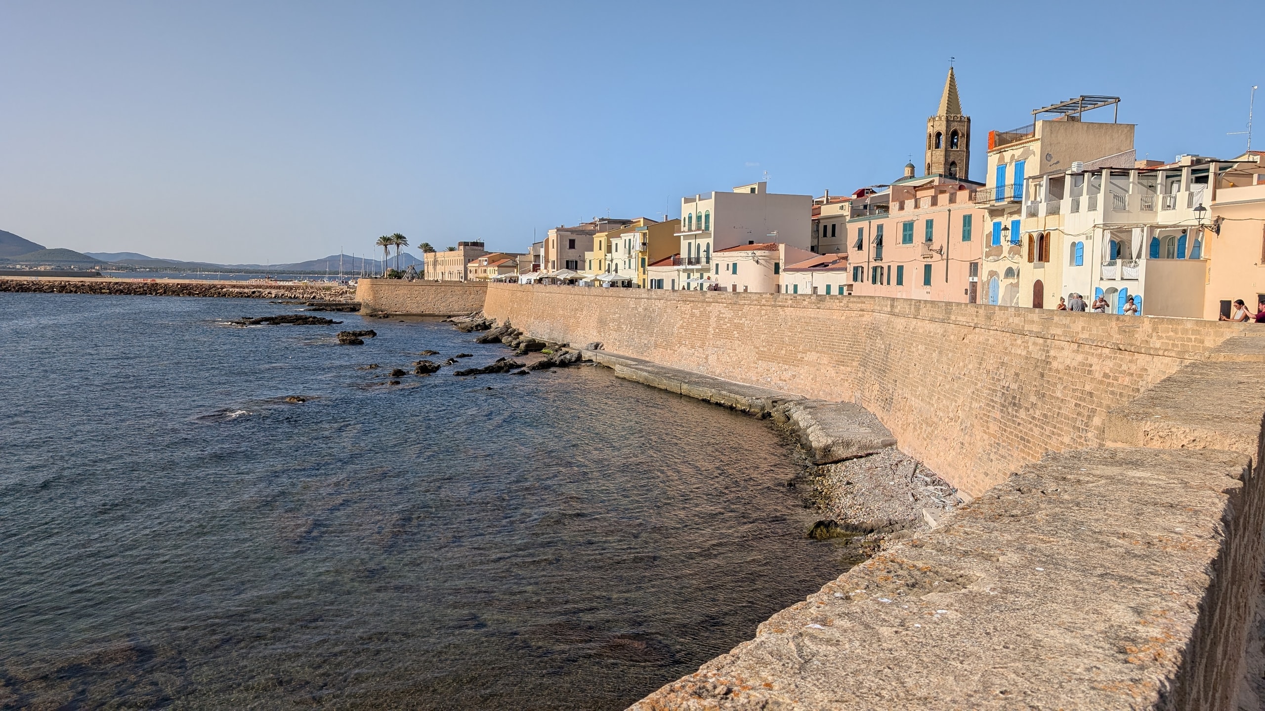 Alghero’s stone sea walls and pastel harbour buildings with the cathedral bell tower under late-afternoon light