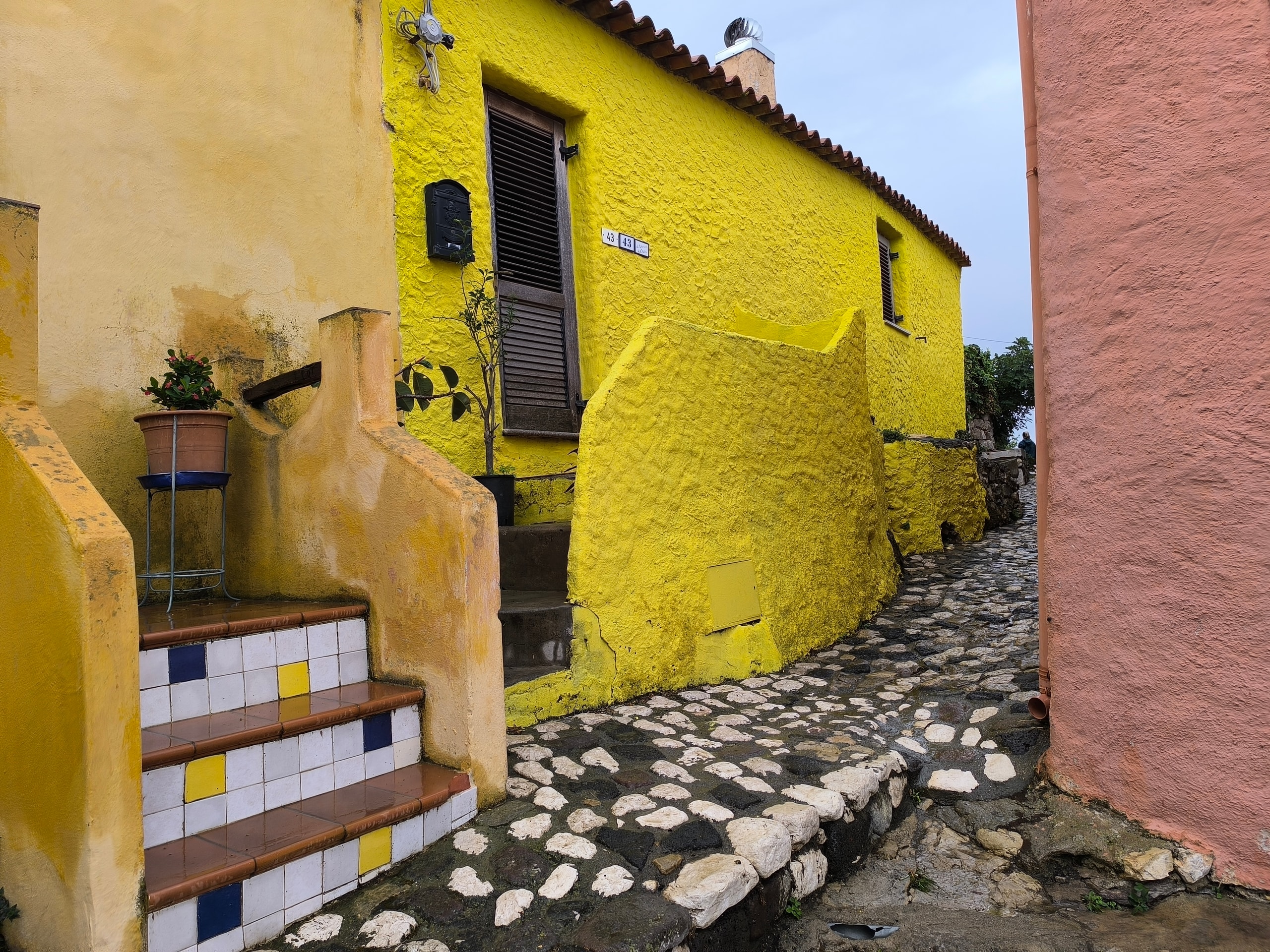 A narrow cobbled lane in Castelsardo with yellow and pink plaster walls, tiled stair risers, and house number 43