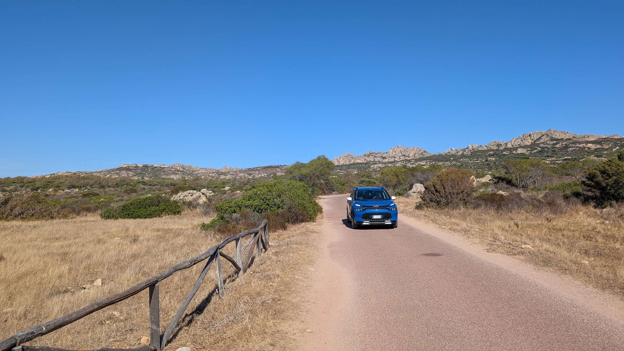 A blue compact SUV on a narrow paved road through dry grass and granite hills in Gallura, North Sardinia