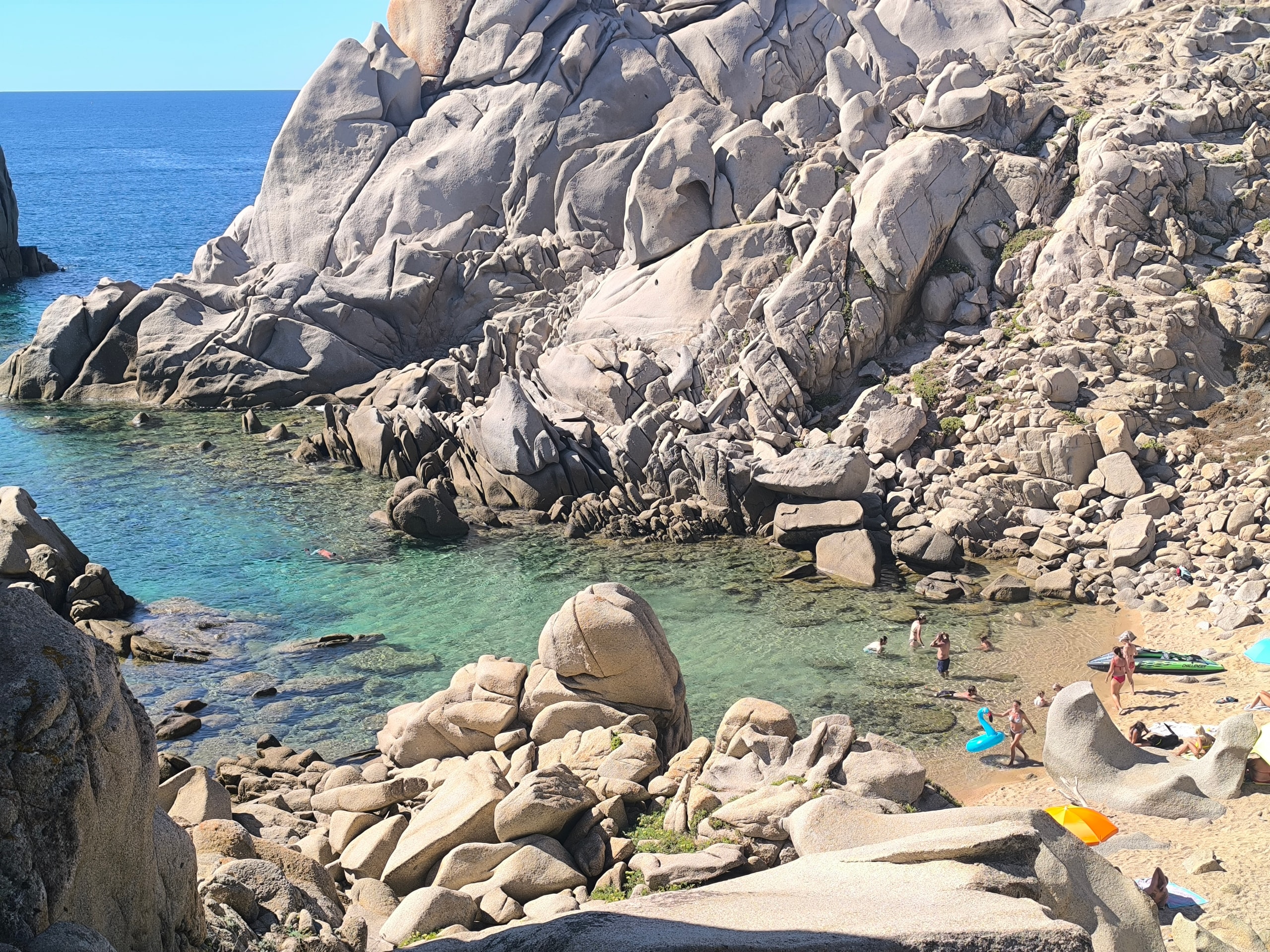 People swimming in a turquoise granite cove at Valle della Luna near Capo Testa, with umbrellas on a small sandy beach