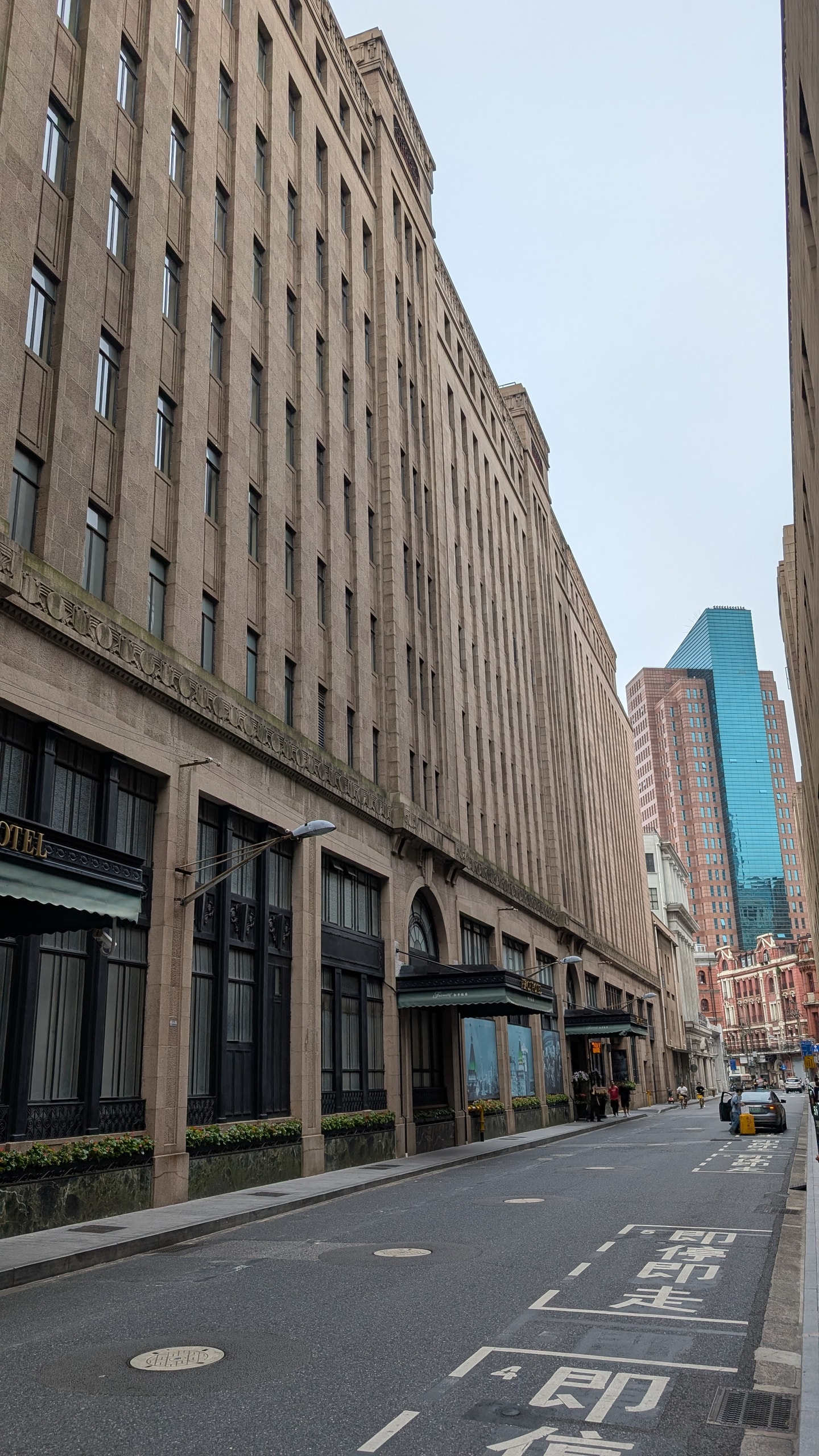 An empty street behind the Bund lined with a long stone office building with green awnings, a blue glass tower rising at the end