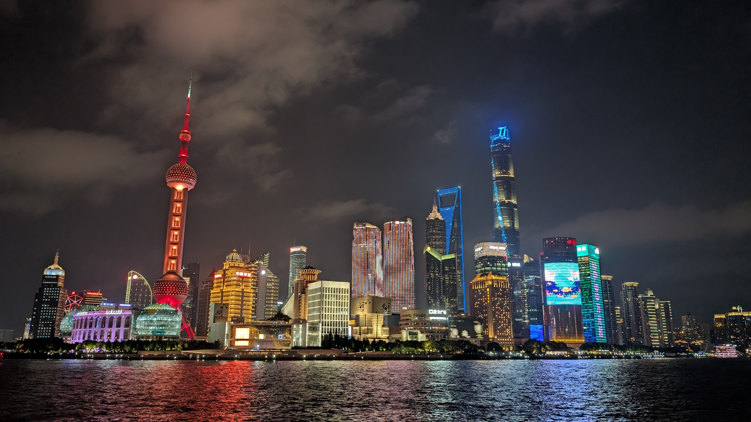 The Pudong skyline lit at night across the Huangpu River, with the Oriental Pearl Tower in red and the Shanghai Tower glowing blue