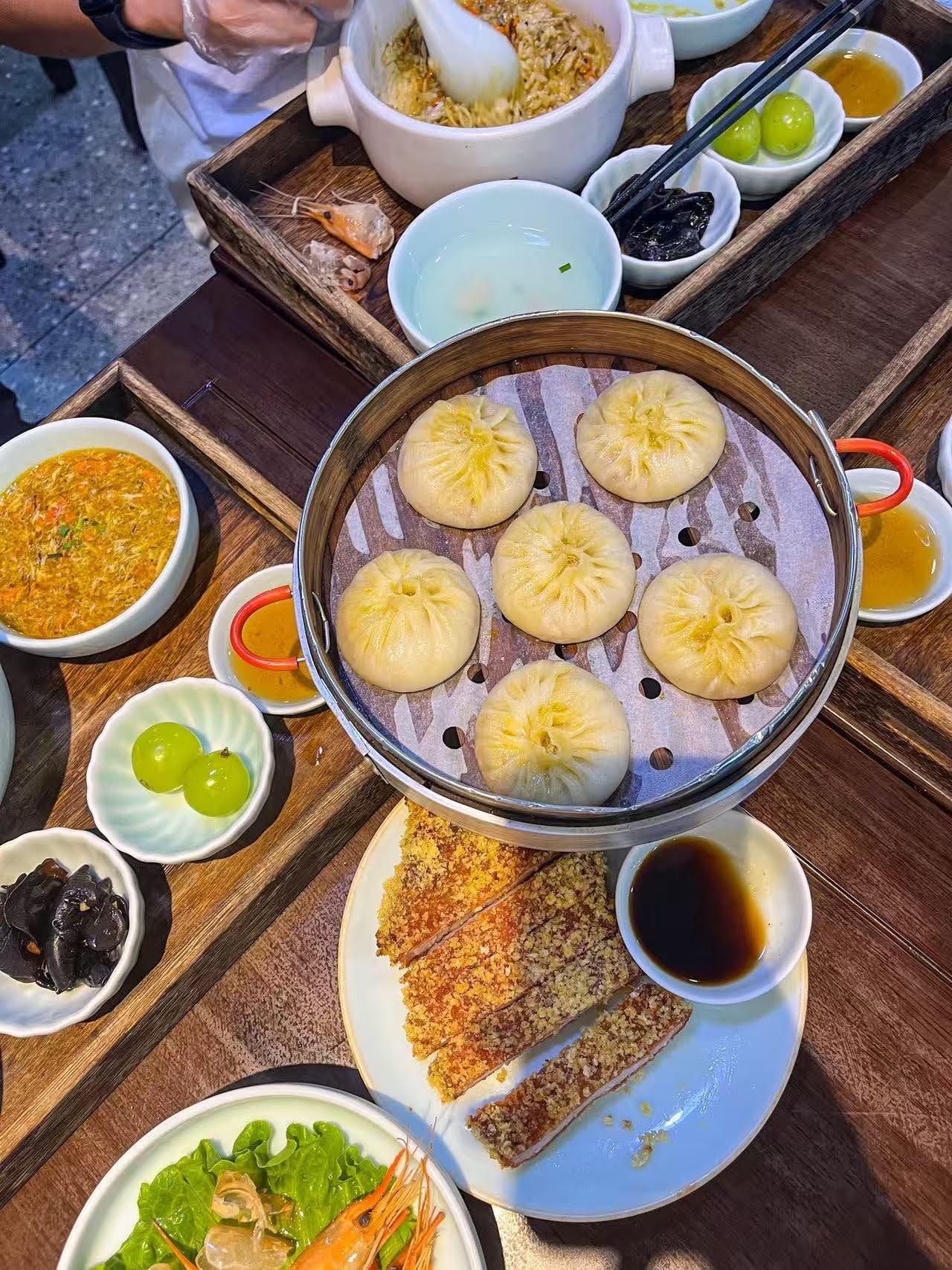 A wooden tray lunch with a pot of crab roe noodles, a steamer of pleated soup dumplings, a sliced breaded pork cutlet with dark dipping sauce, and prawns