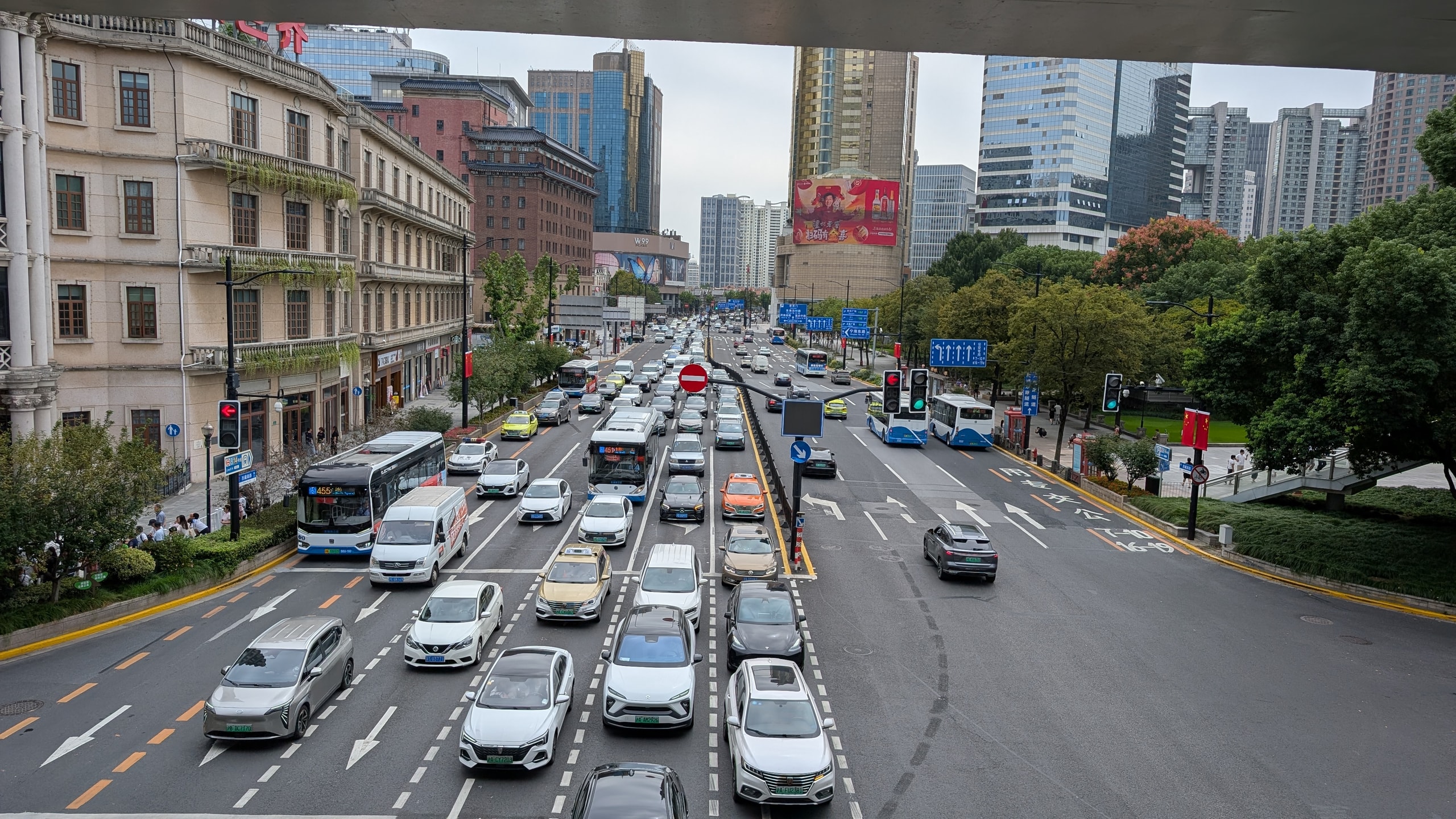 Six lanes of traffic seen from a pedestrian bridge, most cars carrying green electric-vehicle number plates, a bus on the left