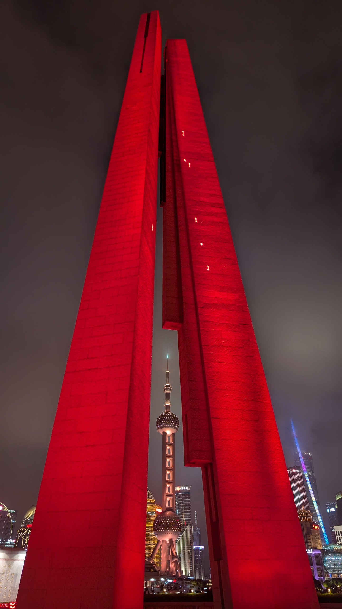 Three tall concrete columns of the Monument to the People's Heroes lit red at night, with the Oriental Pearl Tower framed in the gap between them