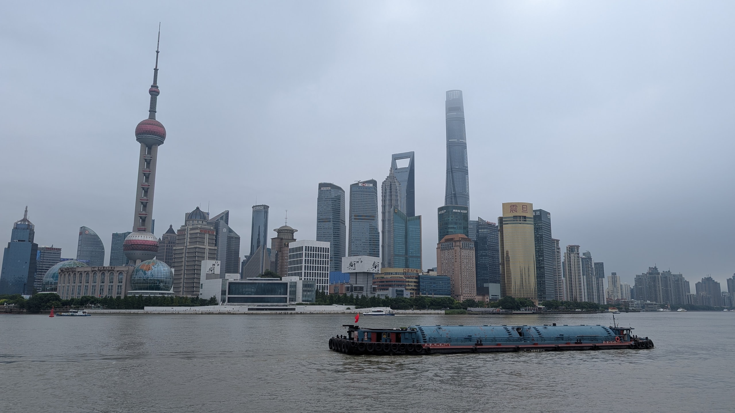 A loaded container barge crossing the grey Huangpu River with the Pudong skyline behind it under low cloud