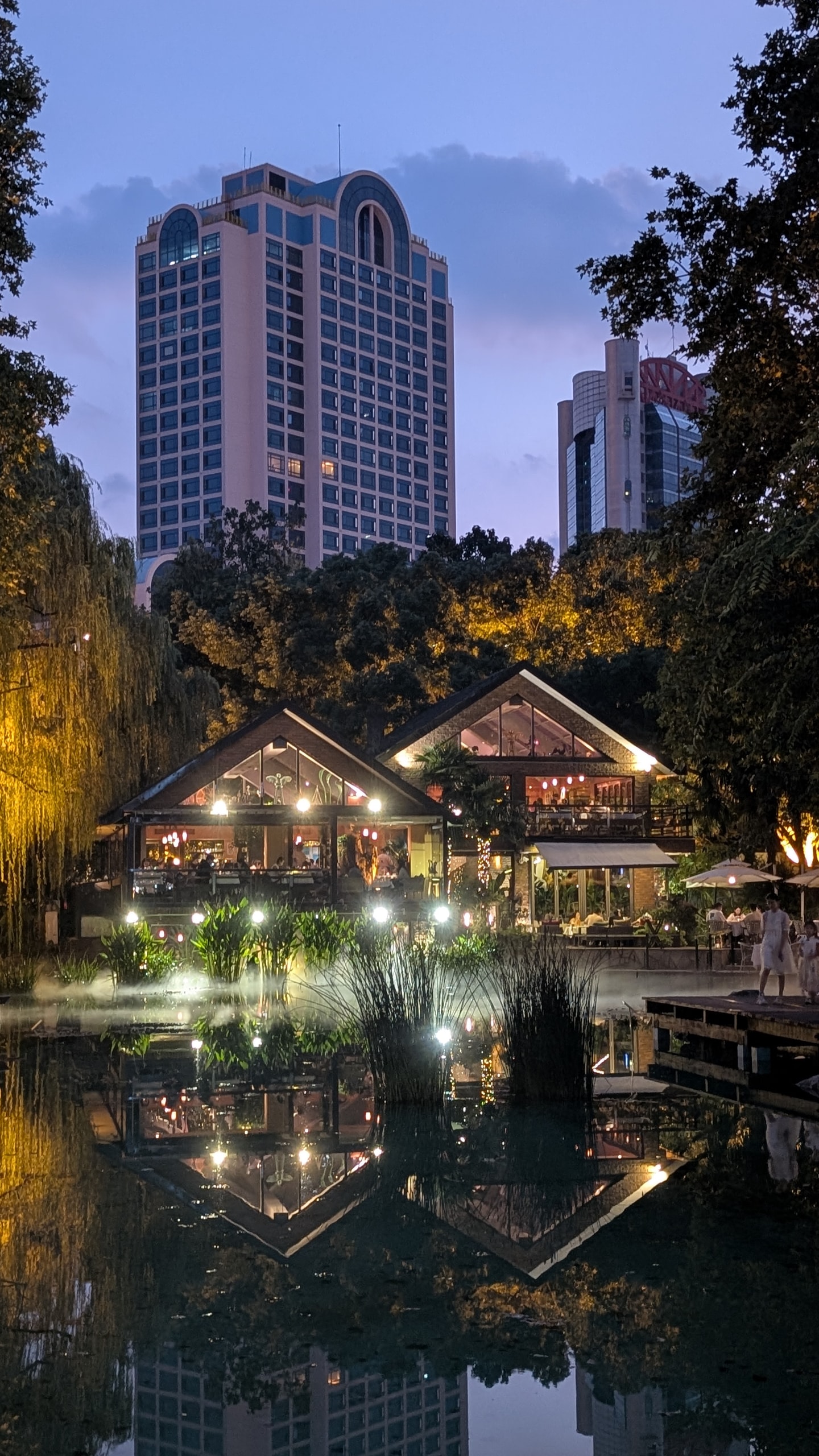 Restaurants with lit glass gables reflected in a park pond at dusk, a tall hotel tower rising behind the trees