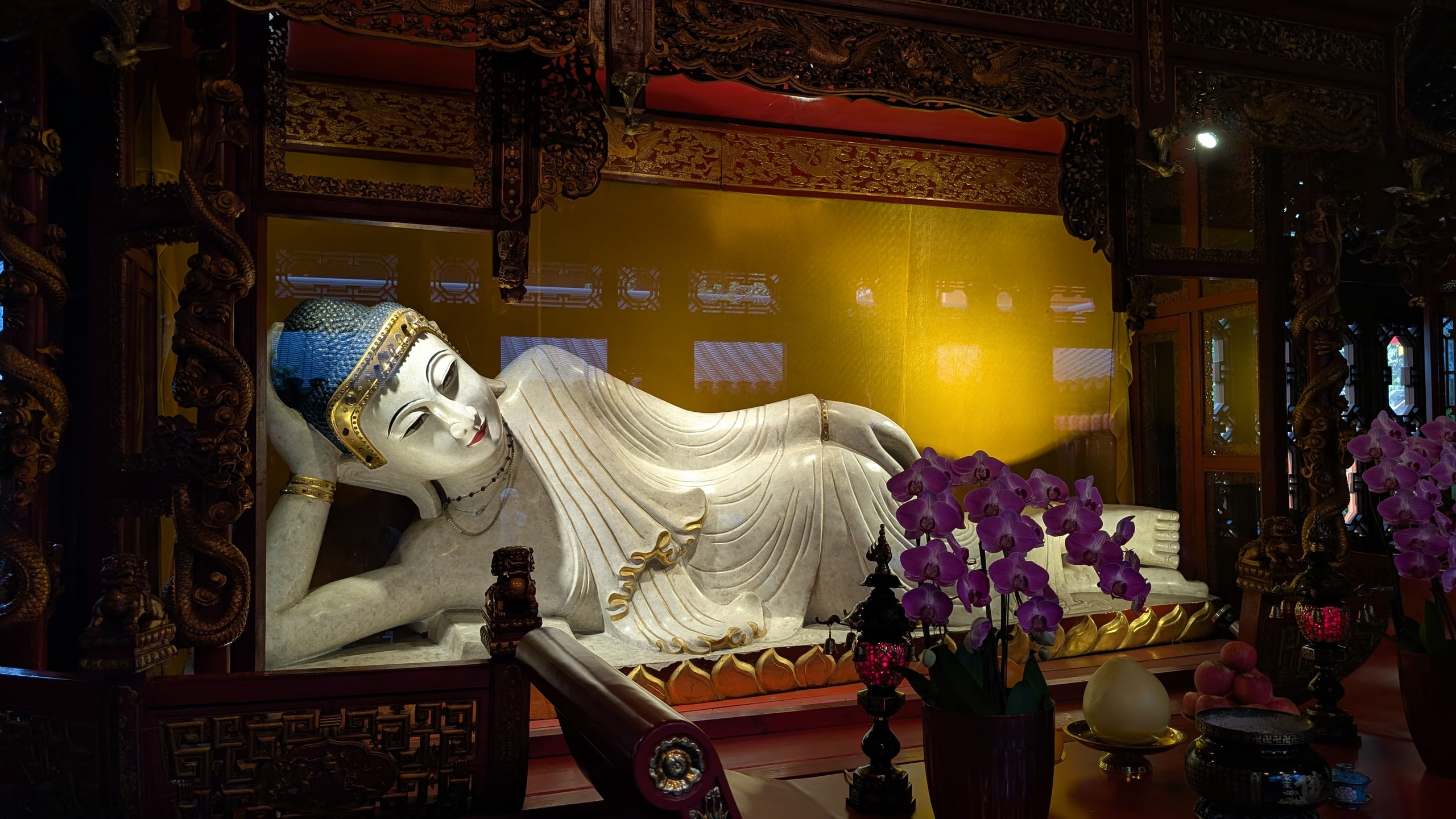 A large white reclining Buddha with gold detailing lying under a yellow wall in a dim temple hall, orchids in front