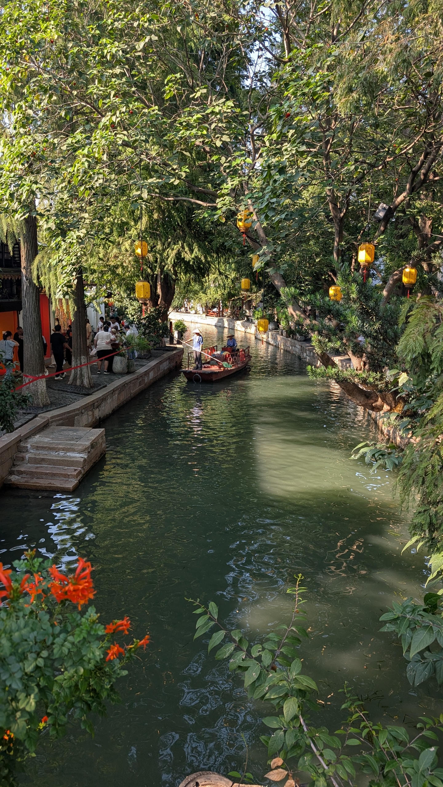 A green canal in Zhujiajiao lined with trees and yellow lanterns, a wooden boat with a standing boatman in the middle