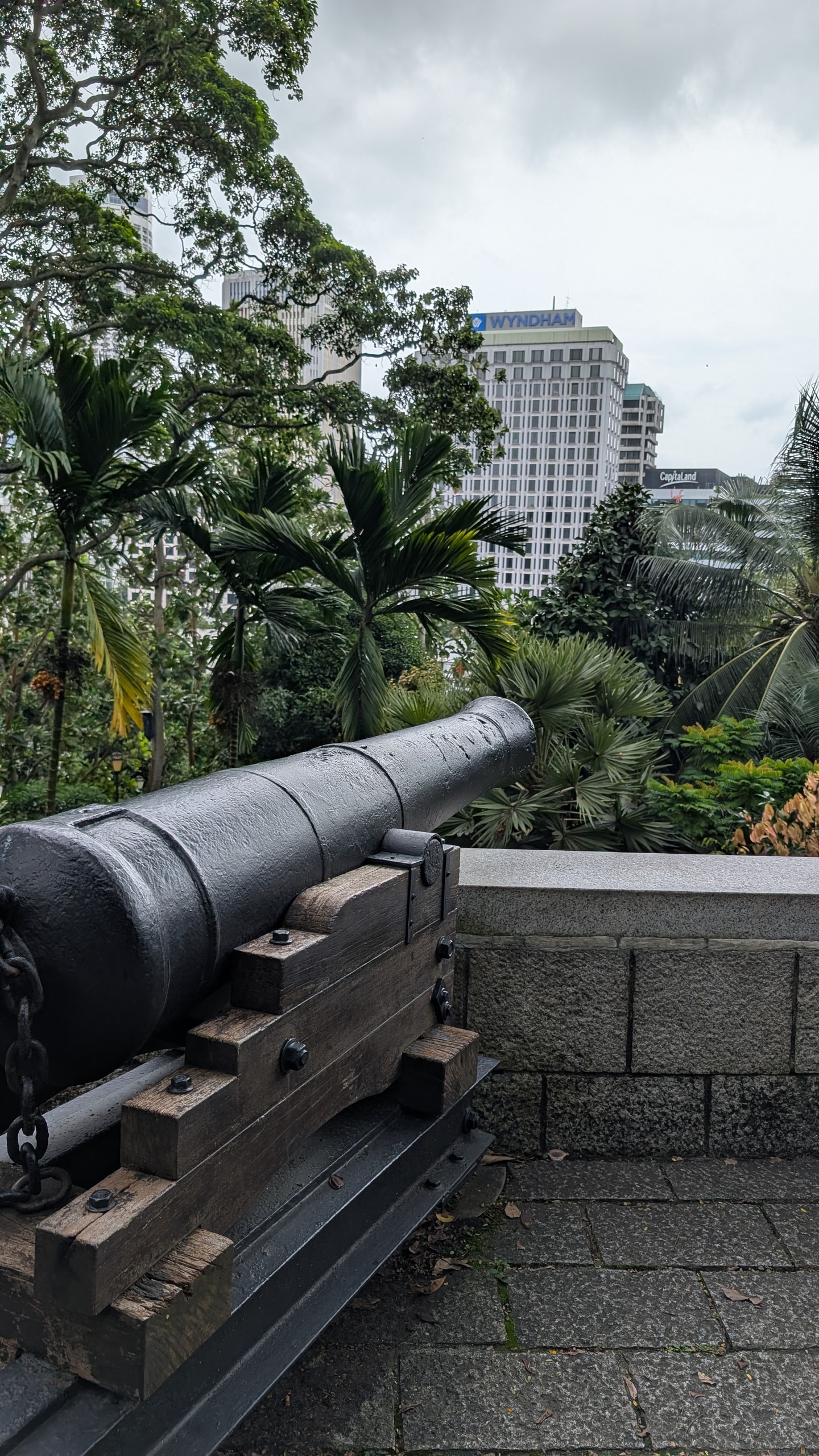 A black nine-pounder cannon on a wooden carriage at Fort Canning, aimed over palm trees toward a white high-rise hotel
