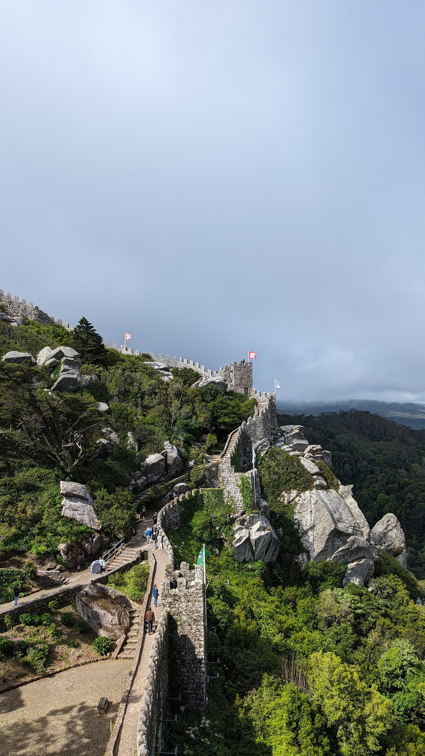 Crenellated stone walls of the Moorish Castle climbing a forested granite ridge in Sintra, with flags on the towers under a cloudy sky