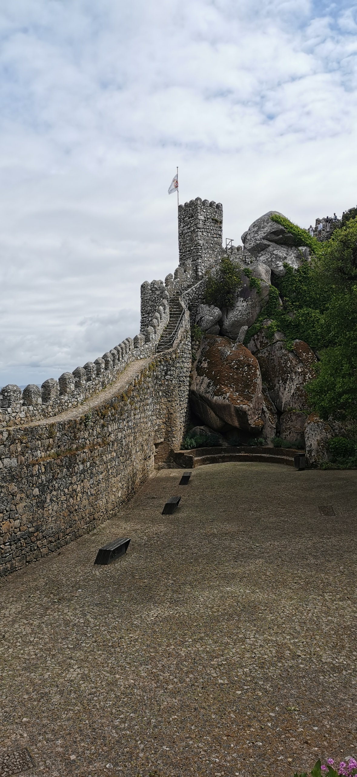 A square battlemented tower of the Moorish Castle with a flag on top, walls built over granite boulders above a gravel courtyard