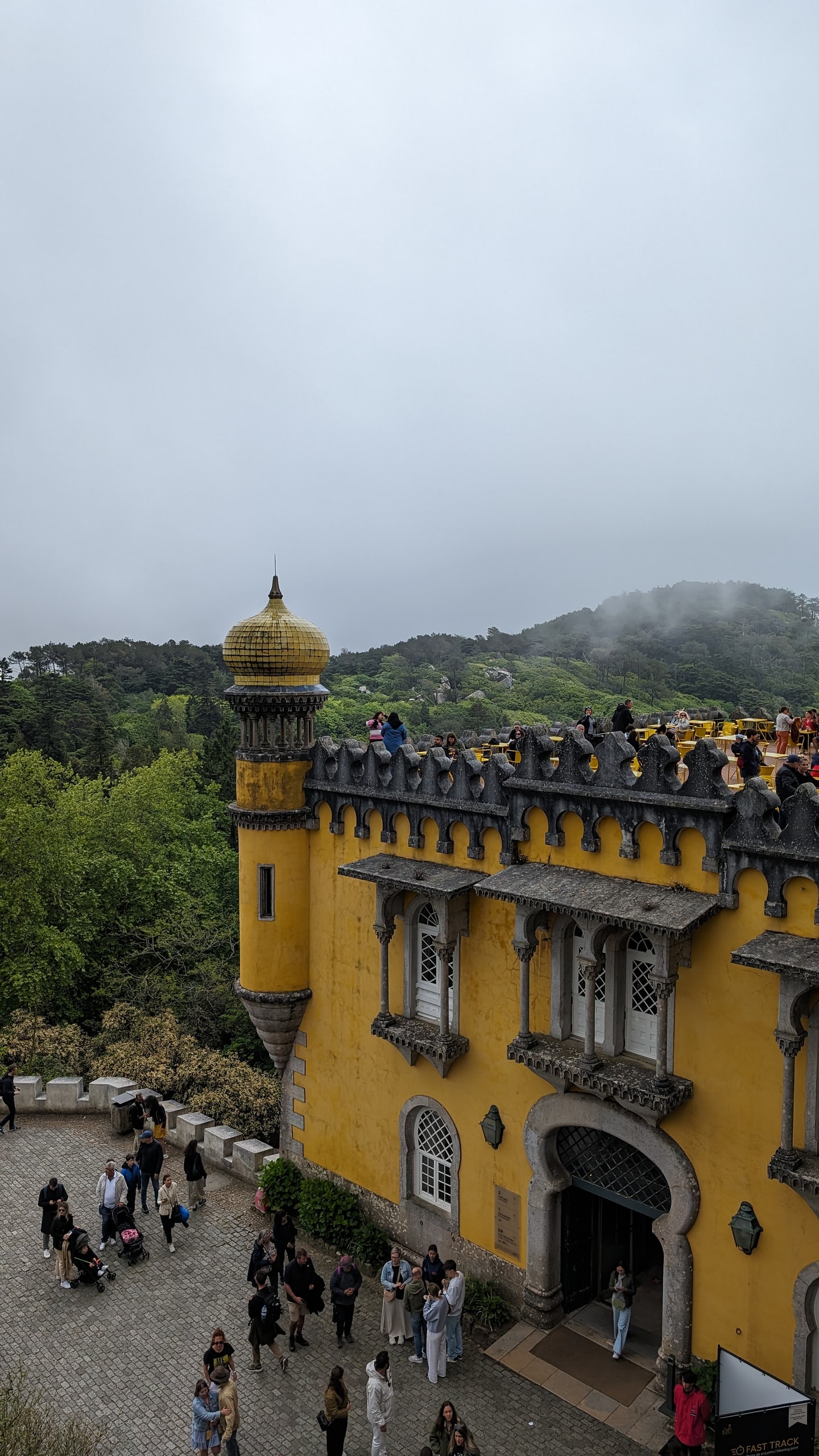 Yellow turret and onion dome of Pena Palace above a cobbled courtyard, with forested hills fading into low cloud