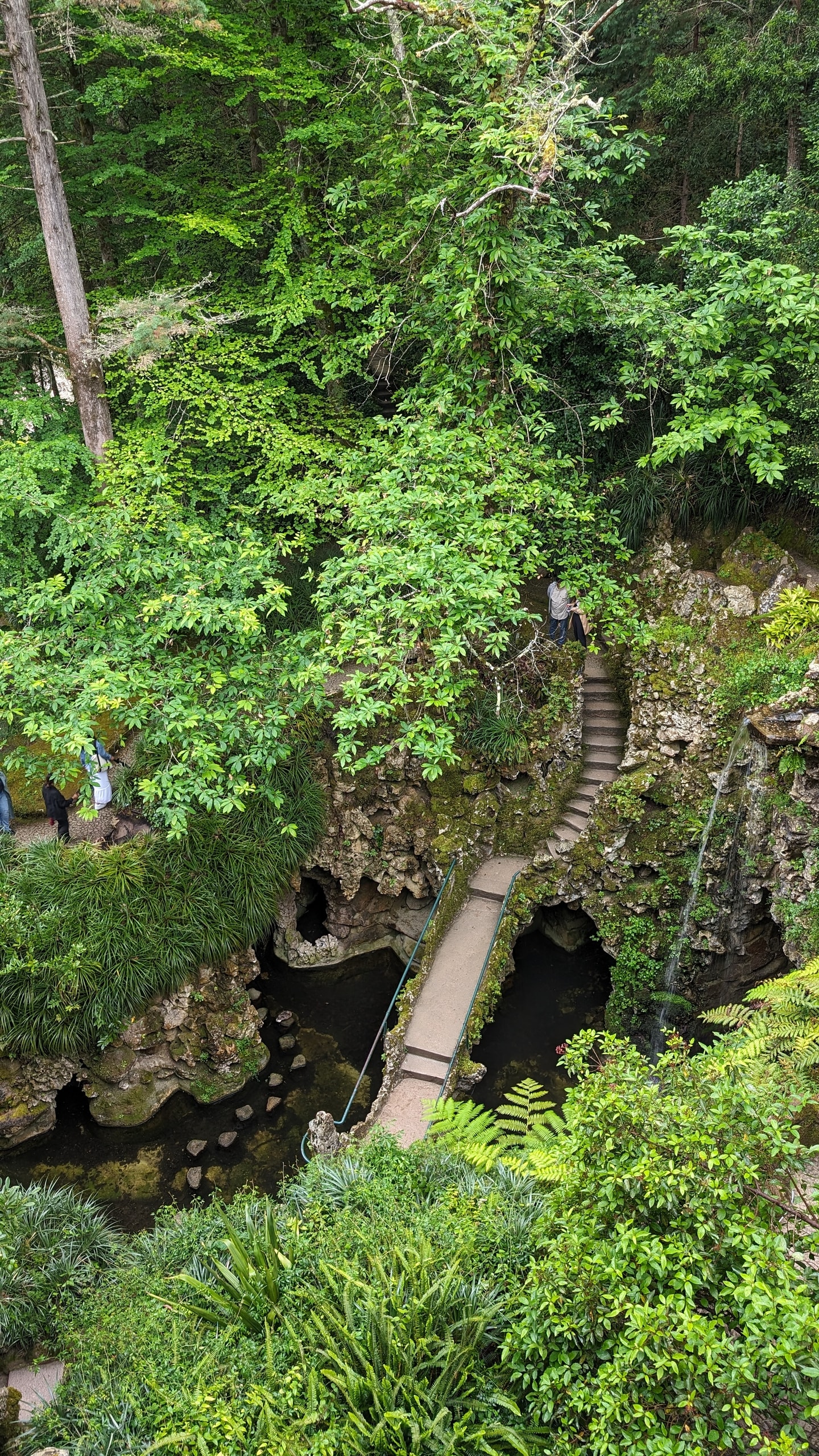 A mossy grotto at Quinta da Regaleira with a stone bridge, circular stepping stones across dark water, and a thin waterfall